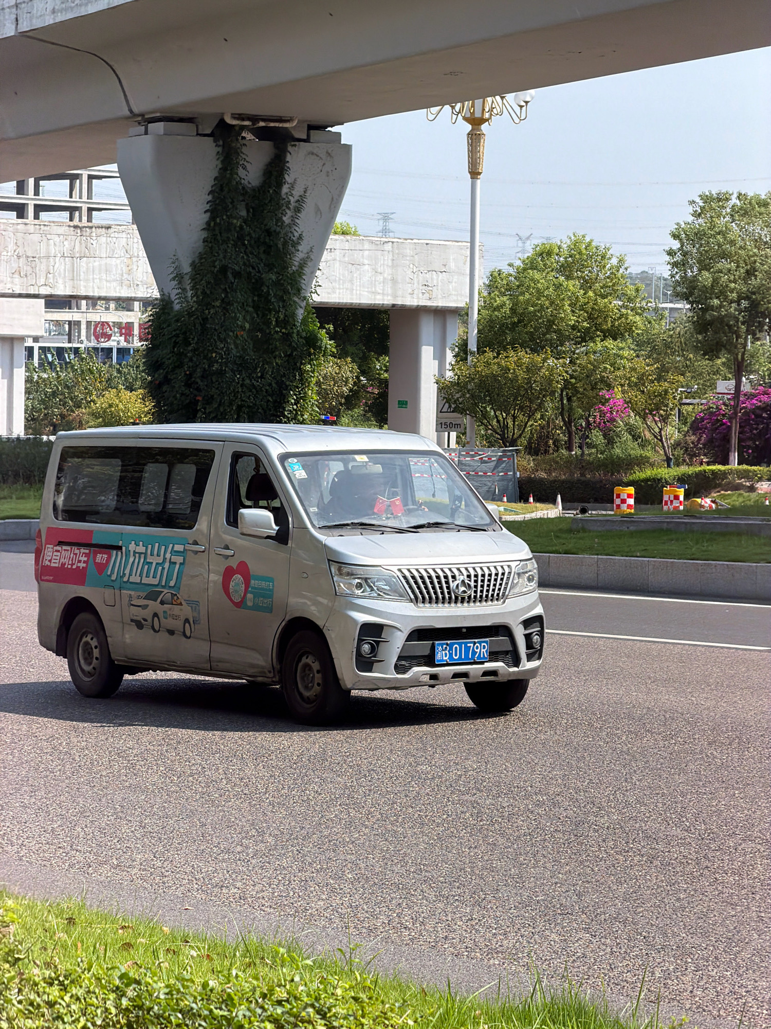 A silver minivan with Chinese characters and graphics is driving on a paved road in Jiangjin, China, under an elevated structure. The minivan has a blue license plate that reads "B-0179R". On the side of the minivan, there are graphics that appear to be advertisements or logos, including a red heart symbol and images of cars. The elevated structure is supported by large concrete pillars, one of which is partially covered in green vines. In the background, there are trees, power lines, and more concrete structures, suggesting an urban or semi-urban environment. The sky is clear and blue. There are traffic cones and barriers visible on the grassy area next to the road.