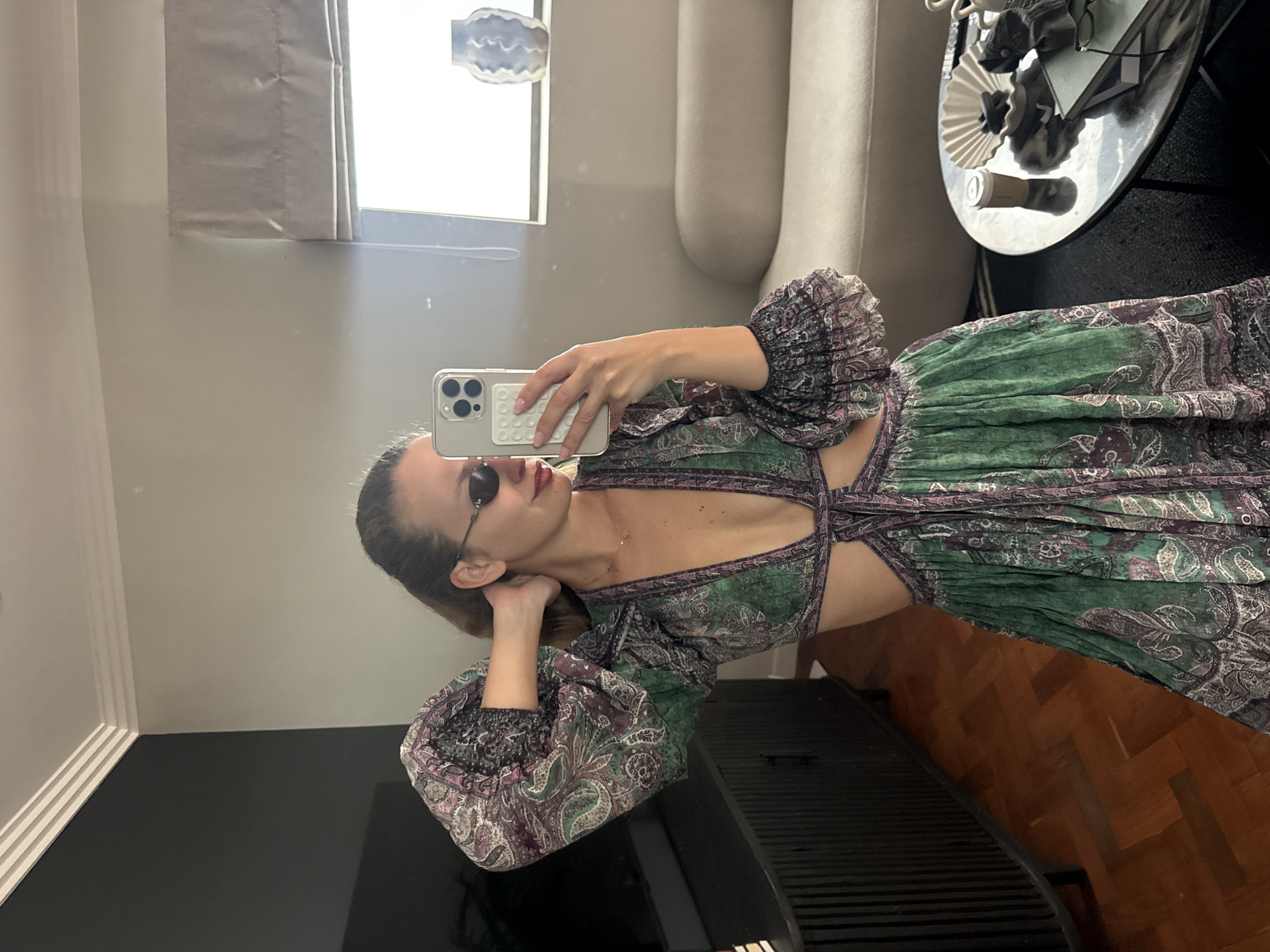 A woman is taking a selfie in a room. She is wearing sunglasses and a green and purple patterned dress with cutouts at the waist. She is holding a smartphone in her right hand, presumably to capture the image. The room contains a window with a view of the outside, a sofa, a table with various items on it, and a large black object, possibly a television, in the foreground. The floor transitions from a dark surface to a wooden parquet pattern. It is not possible to ascertain the location in Cape Town, South Africa.