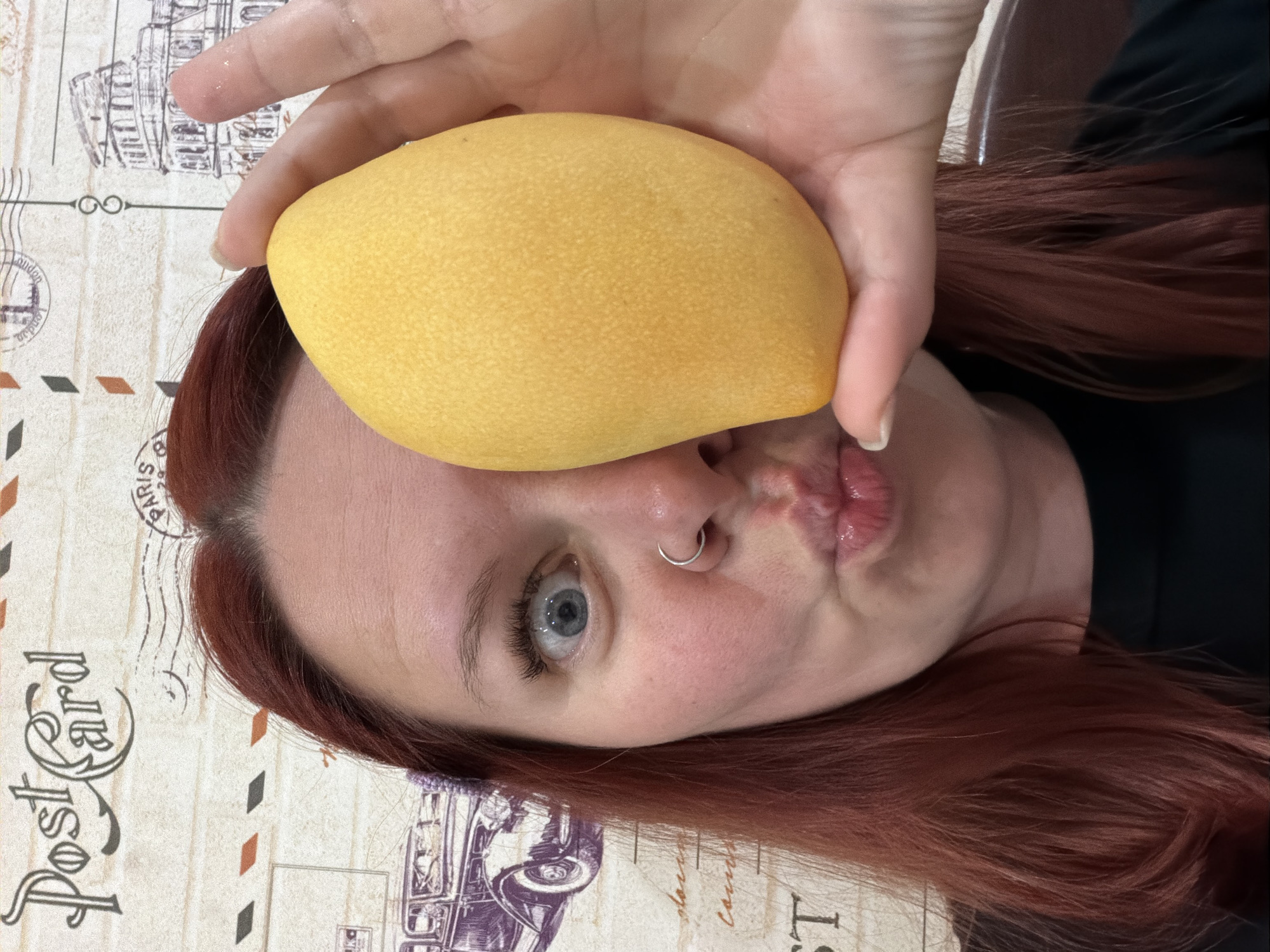 A person with reddish-brown hair is holding a yellow mango over their forehead, making a puckered face. The person has fair skin, blue eyes, and a silver nose ring. The background is a postcard-themed table covering with images of envelopes, cars, and text. The image is rotated counter-clockwise. There is no indication of the city of Chaikovsky, Russia in the image.