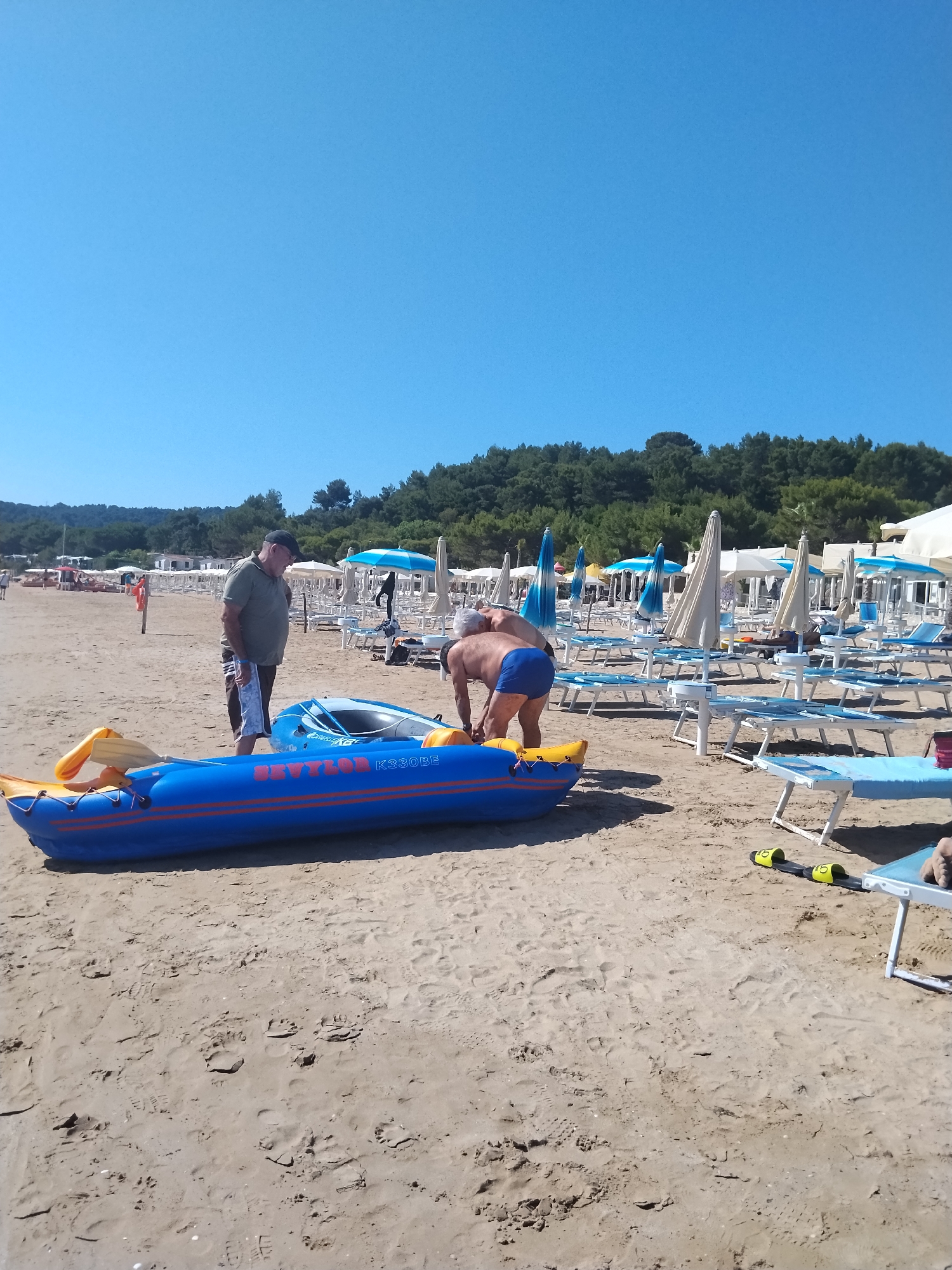 This photograph captures a sunny day on a sandy beach in Peschici, Italy. Two men are the primary subjects, engaged in preparing an inflatable kayak for use. The kayak is bright blue with orange accents and has "SEVYLOR K330BE" printed on its side. One man, wearing a green t-shirt, cap, and patterned shorts, stands looking at the kayak. The other man, wearing blue swim shorts, is bent over, appearing to adjust something on the kayak.
The beach is lined with numerous lounge chairs and umbrellas, indicating a popular tourist destination. The sand is a light tan, with some footprints visible. In the background, a line of trees forms a dense green backdrop against a clear, bright blue sky, suggesting a warm and fair-weather day. The overall scene conveys a relaxed and leisurely atmosphere, typical of a summer vacation by the sea. The time of day is clearly daytime, with the bright sun casting distinct shadows.