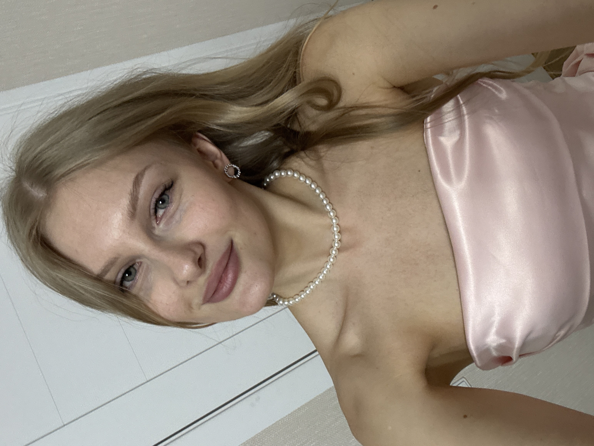 A portrait of a fair-skinned blonde woman with blue eyes, wearing a pearl necklace and a pink satin strapless dress, possibly indoors against a neutral-toned wall and a white door with horizontal lines. No location information is discernible from the image.