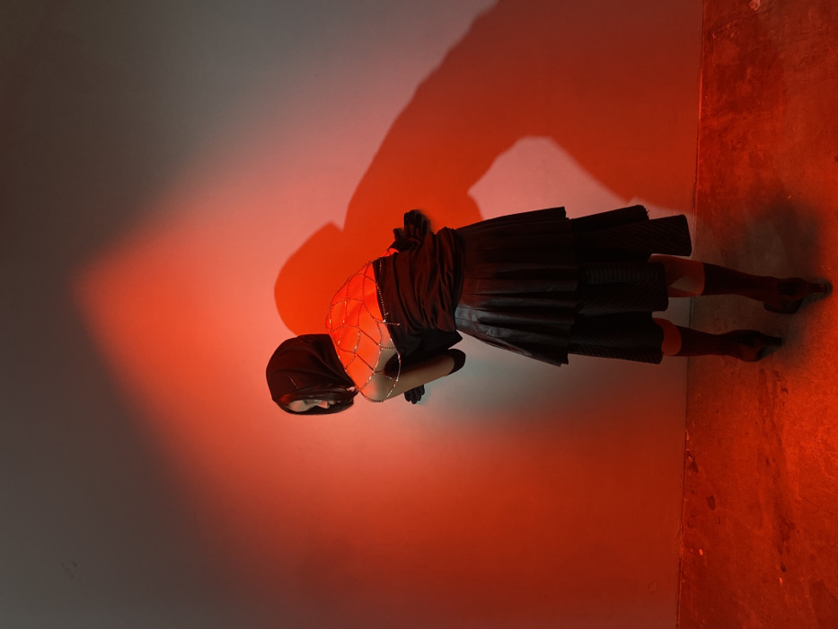 A single individual, appearing female, is depicted indoors, facing away from the camera and turning her head to her left. She wears a dark hooded garment, a dark strapless top or dress, a decorative chain or rhinestone harness forming a web-like pattern across her upper back, long black gloves, a black tiered pleated skirt, black socks or hosiery, and black high-heeled shoes. Her body is angled to the left, with her left arm slightly bent and hand near the wall. The subject's dark shadow is prominently cast on the plain, light-colored wall to her right. The scene is illuminated by strong red light from a low angle, casting a deep red hue on the lower wall and the floor, which appears to be a textured, possibly concrete surface. The upper wall transitions from red to a muted grey-white. There are no identifiable architectural features, street signs, or geographical markers within the image to determine a specific location within London, United Kingdom.