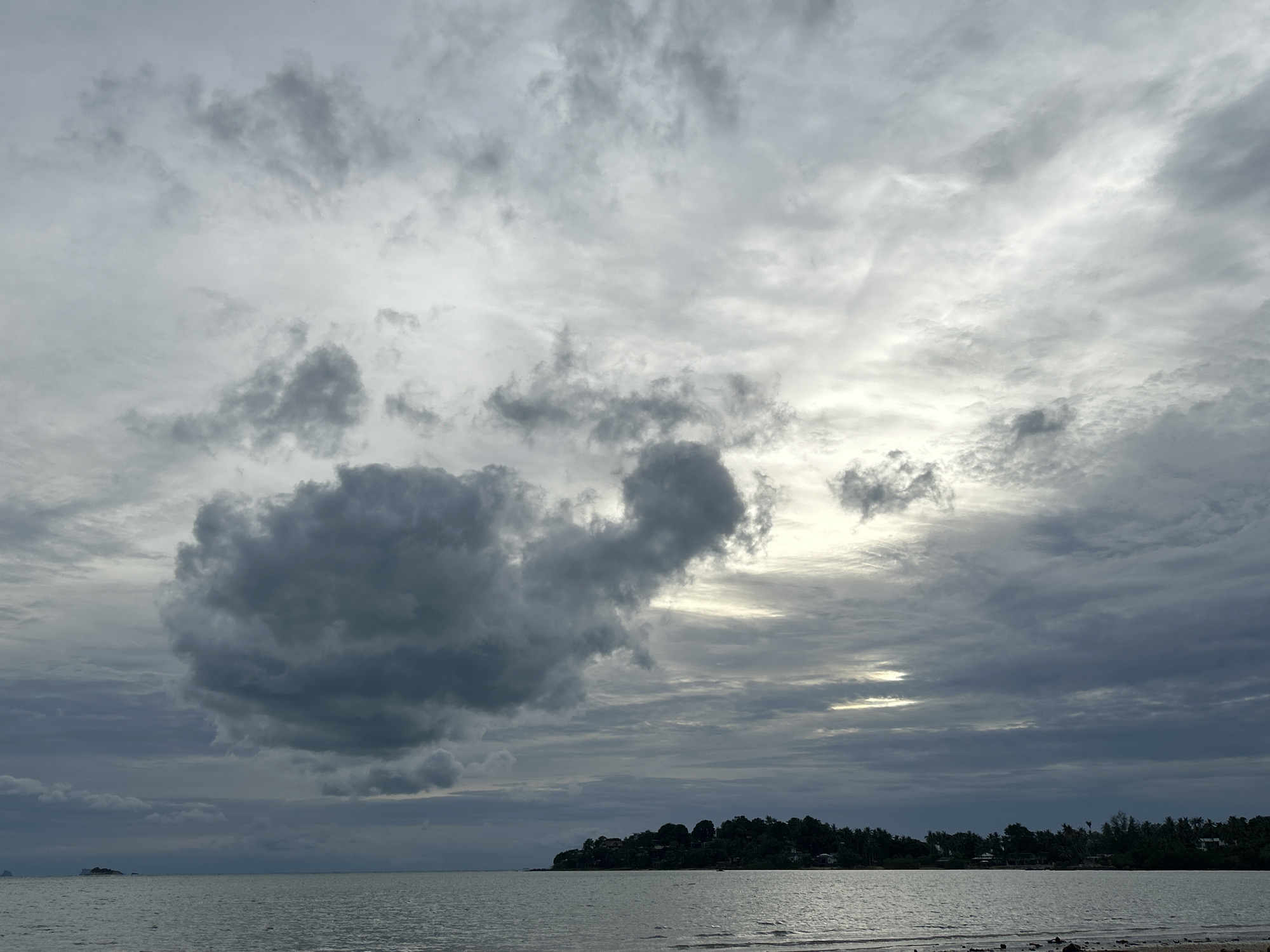 This image shows a cloudy sky over a body of water, likely the ocean, with a treed coastline and some buildings visible on the land. A large, dark cloud is prominent in the upper left of the frame. The water is calm, with a slight ripple. The shoreline is composed of sand and small stones. The overall scene suggests a slightly overcast day in Ko Pha-ngan District, Thailand, with a view of the coast. There are no people or animals visible in the image.
