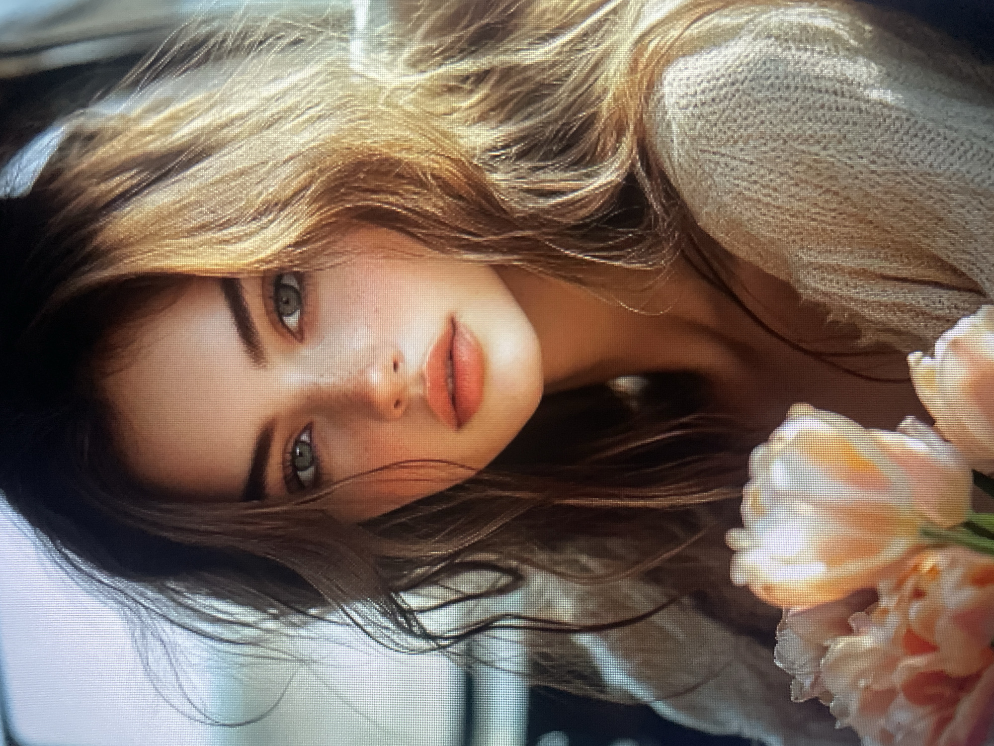A young woman with long, wavy, light brown hair is lying down, with her head tilted towards the viewer. Her eyes are a striking green, and she has a light dusting of freckles across her nose and cheeks. Her lips are full and painted a soft coral shade. She is wearing a light-colored, textured sweater. In the foreground, to the right of the frame, a bouquet of pale pink and white tulips is partially visible. The scene appears to be indoors, with a blurred background that suggests a soft, ambient light, possibly from a window. The overall mood is serene and intimate, with a focus on the woman's delicate features and the soft textures of her hair, clothing, and the flowers. The image evokes a sense of peacefulness and natural beauty. There is no visible text in the image.