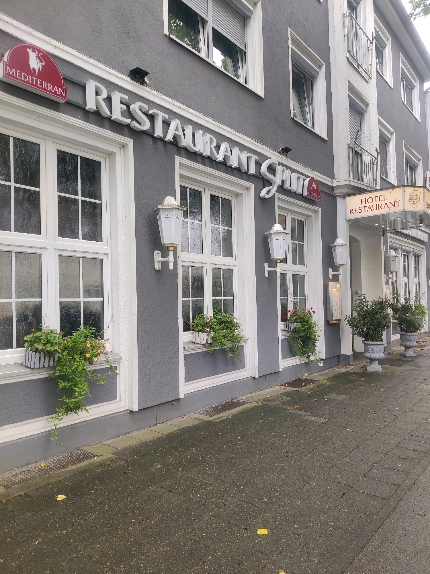 The image shows the exterior of a building located in Emmerich am Rhein, Germany, which houses a hotel and restaurant. The building has a grey facade with white-framed windows. Above the entrance, a sign reads "HOTEL RESTAURANT". Another sign above a window on the left side reads "MEDITERRAN". White lanterns are mounted on the facade next to the windows, and planters with greenery are situated beneath some of the windows. The sidewalk in front of the building is paved with grey tiles, and there are fallen leaves on the ground, suggesting an outdoor setting. The overall scene depicts the street-facing facade of a commercial establishment.