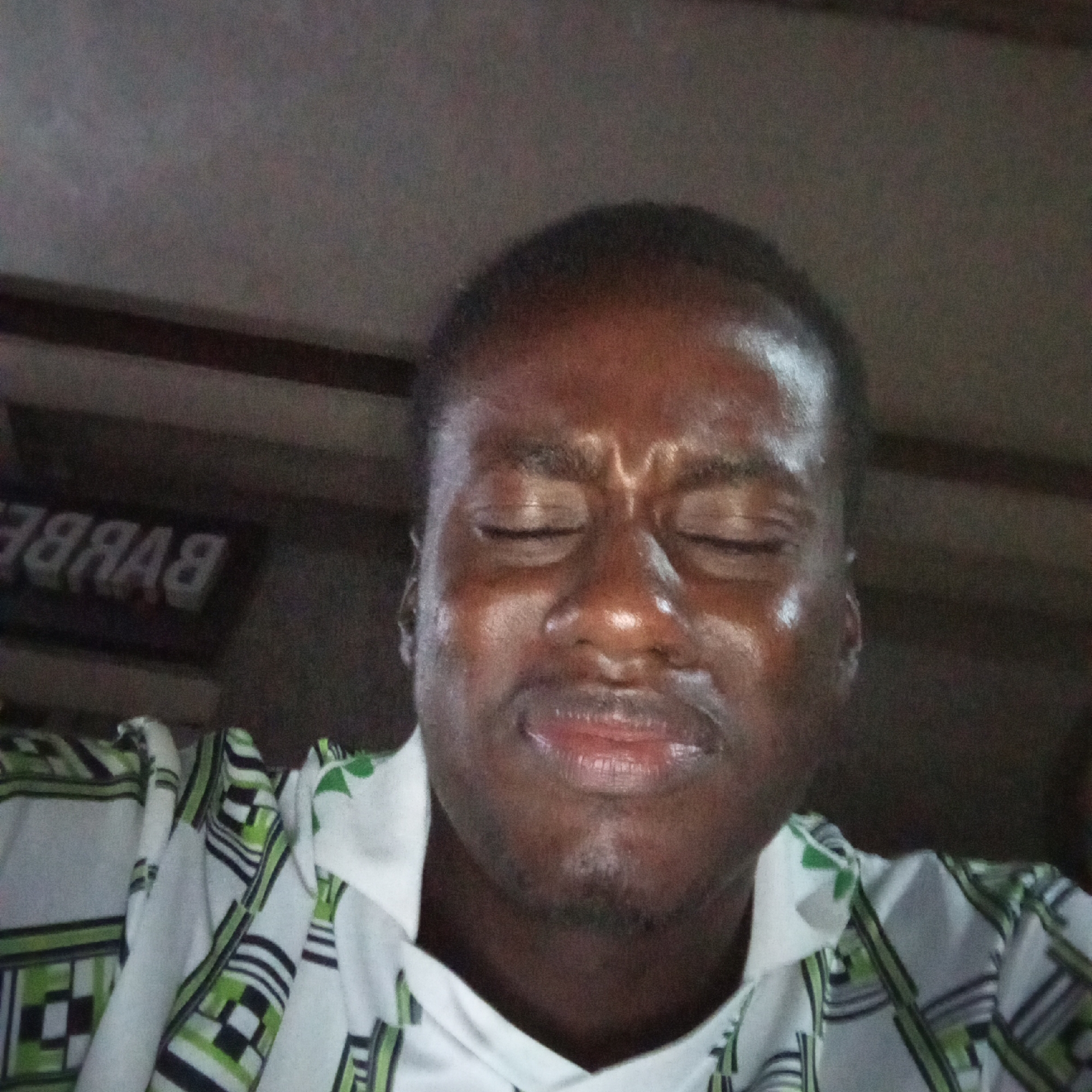 A man with closed eyes, his face appears sweaty, is shown in a close-up shot. He is wearing a white shirt with a green and black geometric pattern. In the background, a sign with white letters on a dark background is visible, reading "BARBER" in reverse. The overall lighting suggests an indoor setting. The image quality is poor, with visible noise and blur. This image was taken in Enugu, Nigeria.