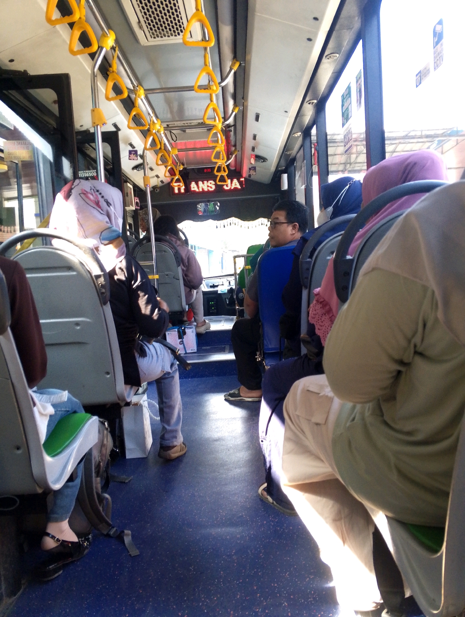 The image shows the interior of a public bus in Meri, Indonesia. The bus is crowded with passengers. In the foreground, a person wearing a light-colored hijab is seated on the right, facing away from the camera. To their left, another person is seated, wearing a blue hijab and a white face mask. Further to the left, a man with glasses wearing a blue shirt is visible. Several rows of seats are occupied by passengers. Yellow handrails are suspended from the ceiling. The floor of the bus is blue. The bus appears to be in motion, with sunlight streaming through the windows.