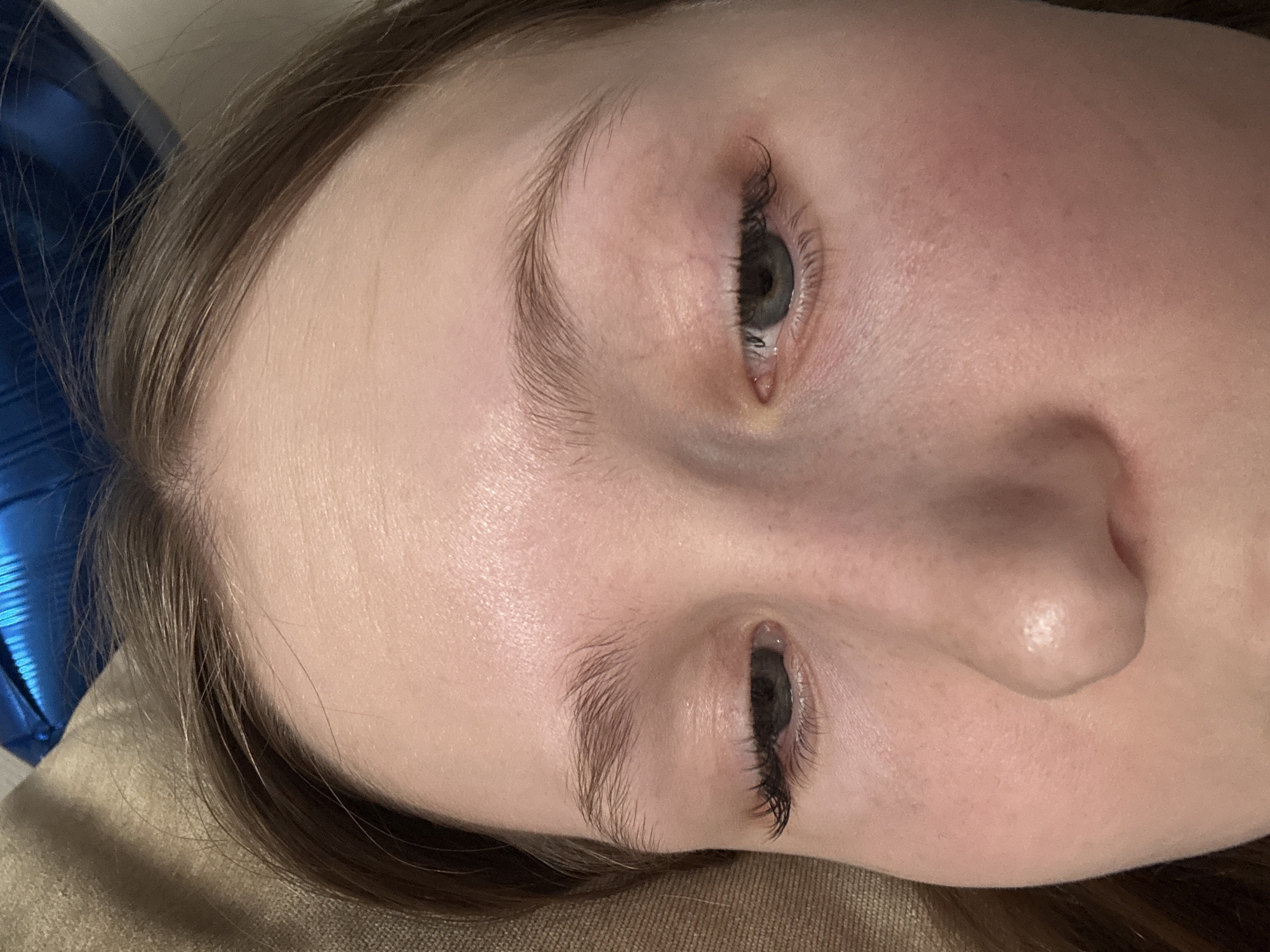 A close-up, high-angle shot of a fair-skinned person's face. The face is tilted slightly to the right, with the nose and right cheek dominating the foreground. Both eyes are visible, with gray irises and dark eyelashes. The eyebrows are thin and light brown. The skin appears smooth and clear with slightly rosy cheeks. Part of the person's light brown hair is visible on the left side of the frame. A blue object, possibly a balloon, is partially visible in the upper left corner. The background is a neutral, beige color, possibly fabric. The city of Chaikovsky, Russia is not visible.