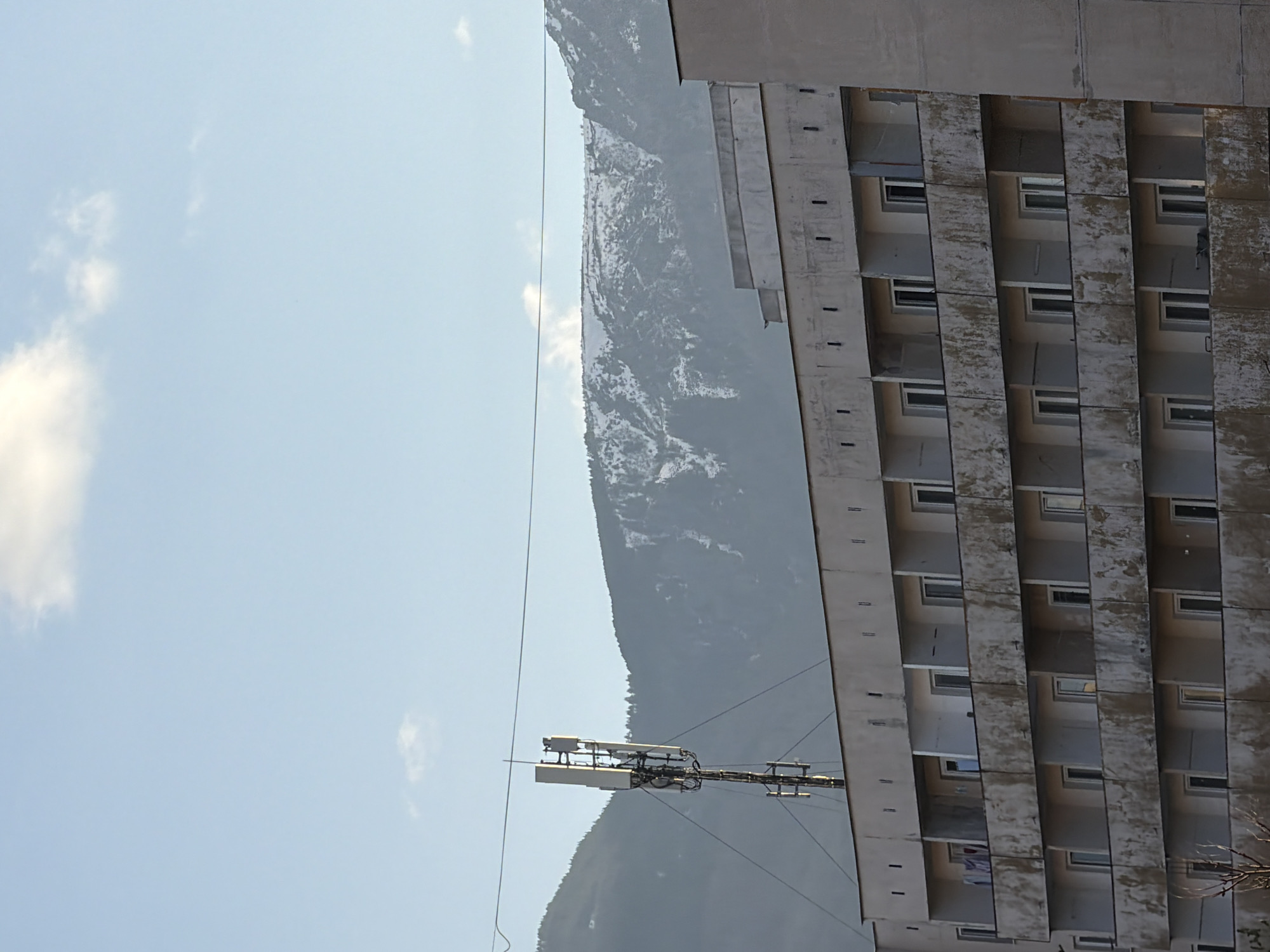 The image shows a tall, gray building with multiple rows of balconies and windows, possibly an apartment complex, with a mountain range in the background. An antenna structure is visible in the foreground. The scene is set against a light blue sky with some clouds. The building appears to be in Sofia, Bulgaria, based on architectural style, landscape and the presence of the mountain range.