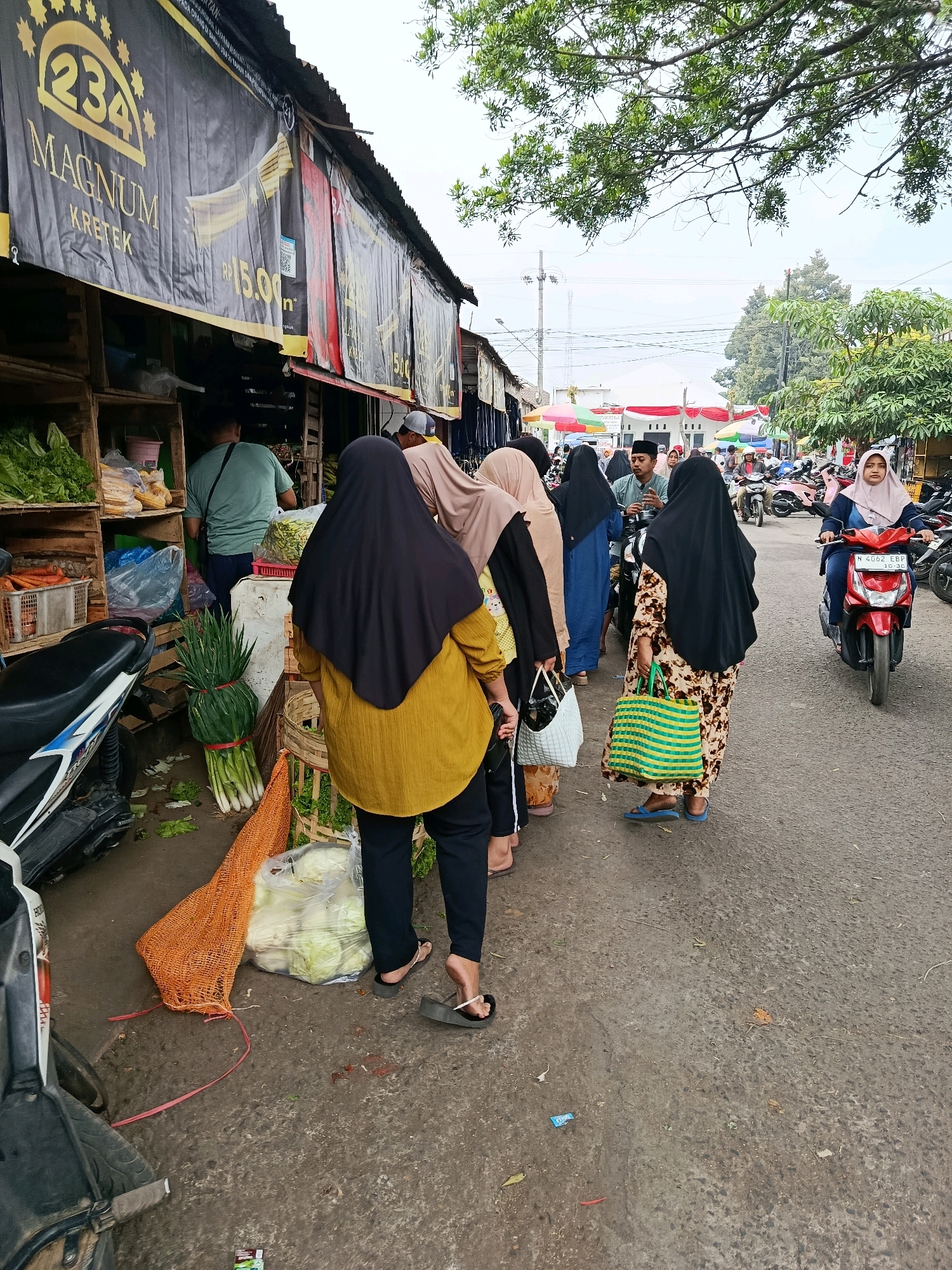 The image captures a street scene in Bululawang, Indonesia, likely a market area, with a focus on a row of stalls. In the foreground, several people, predominantly women wearing hijabs, are walking or standing, some carrying shopping bags. One woman in a yellow top and black pants stands facing away from the camera, next to a display of produce including green onions and cabbages. Another woman in a patterned dress and blue flip-flops walks further down the street carrying a green and yellow striped bag. Behind them, a red motorbike with a rider wearing a light blue hijab is visible, along with other motorbikes parked further in the background. The stalls display various goods, including vegetables. A large black banner with yellow lettering, advertising "234 Magnum Kretek," hangs above one of the stalls. The environment appears to be an outdoor marketplace with a paved road and overhanging trees.