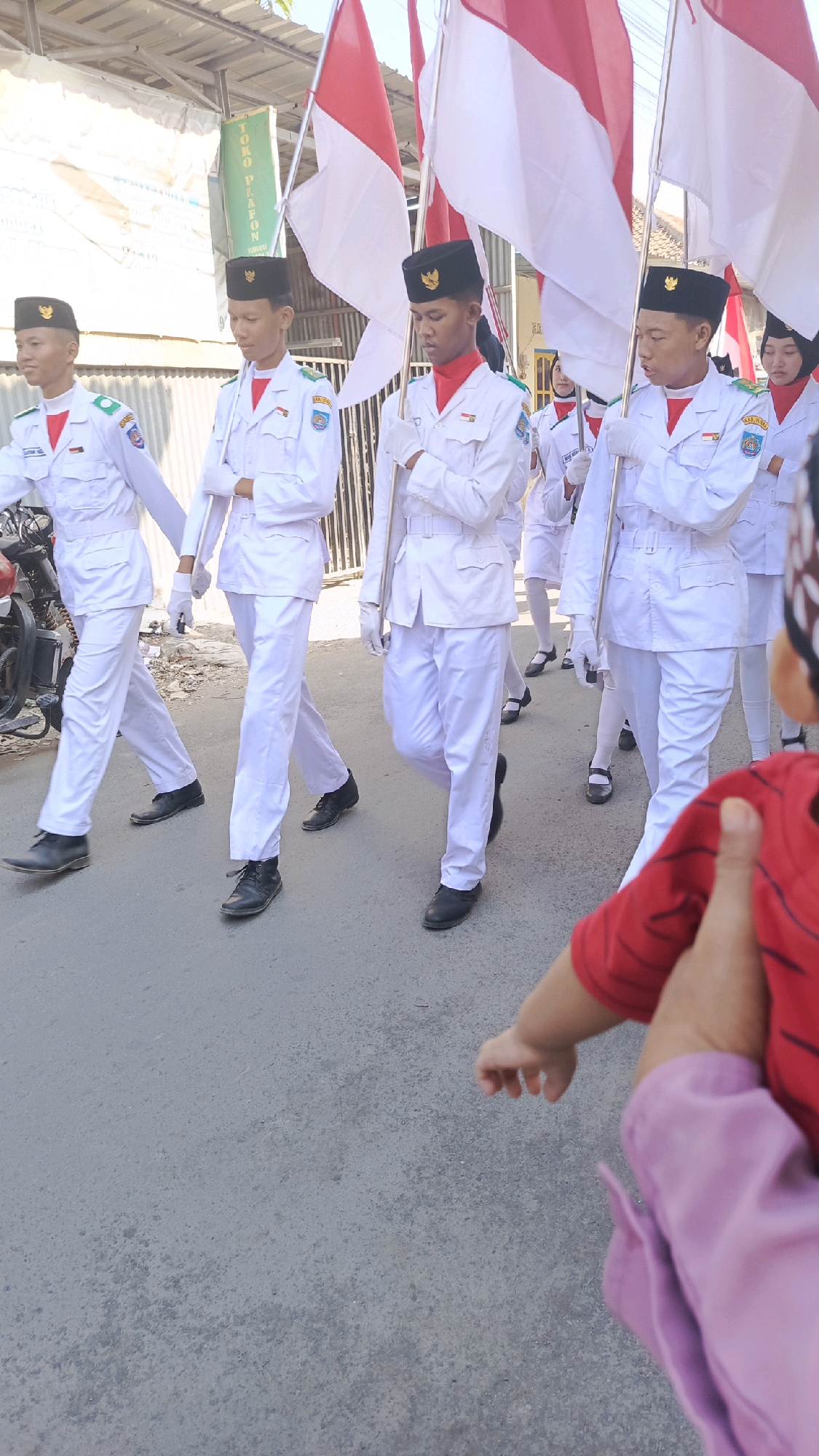 The image depicts a group of young people in white uniforms and black hats marching outdoors on a paved street in Pekauman Kulon, Indonesia. The individuals are holding Indonesian flags, which are red and white. The uniforms are crisp and appear to be ceremonial attire, suggesting a parade or formal event. In the foreground, a child wearing a red shirt and purple garment is being held, with their arm and hand visible. The background shows buildings and a fence. The lighting indicates it is daytime, likely sunny.