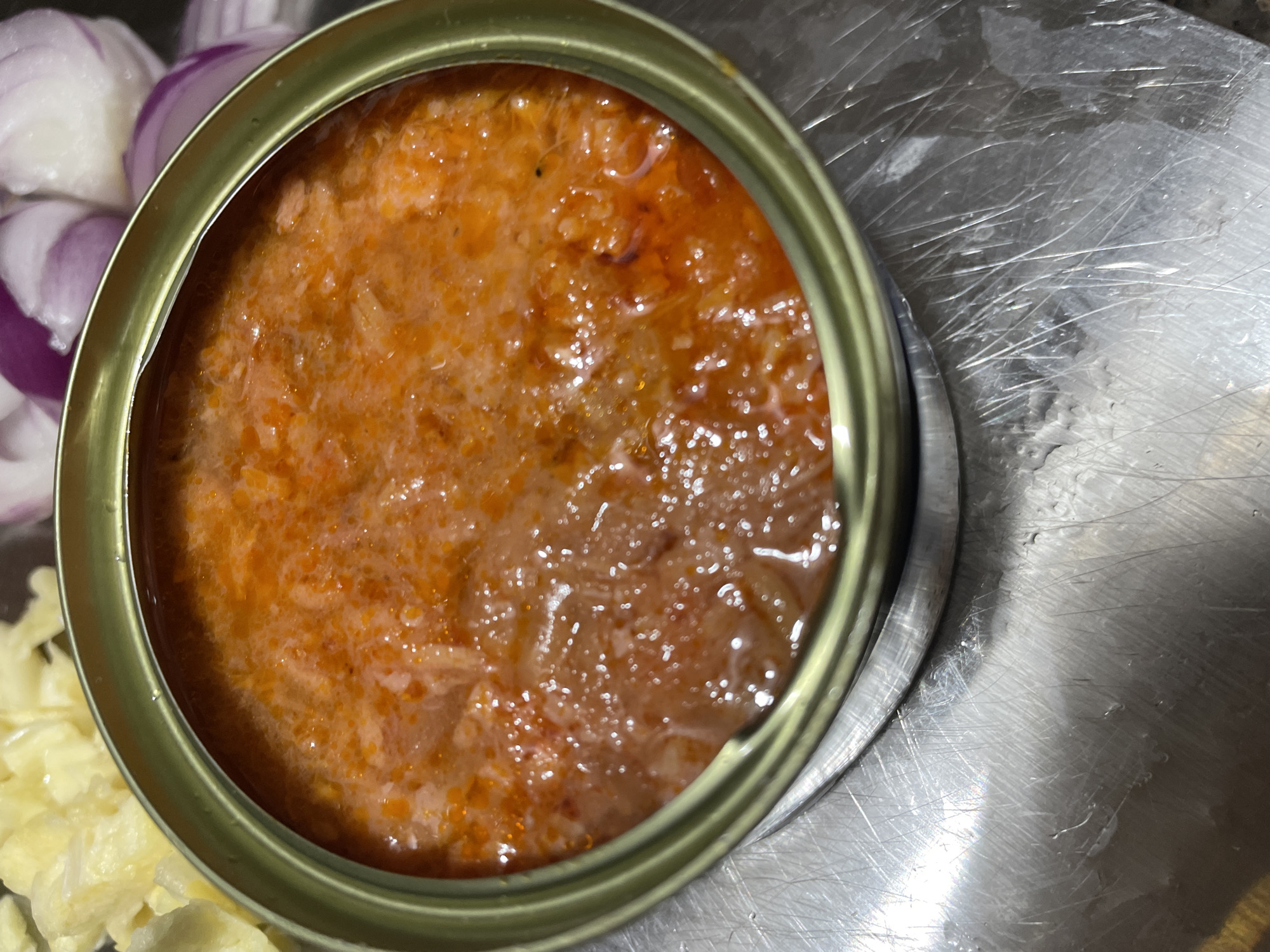 An overhead view captures an open, cylindrical metallic food can, containing a viscous, textured, orange-red substance with suspended particulate matter. The can's rim is a reflective silver-gold tone. The can rests upon a brushed metallic surface characterized by numerous intersecting linear scratch marks and subtle surface reflections. In the blurred background to the upper left, several light yellow, irregularly shaped chunks are visible. To the upper right, out-of-focus segments of purple and white, consistent with chopped red onions, are present. No individuals are depicted, and no direct actions or interactions are occurring beyond the static arrangement of food items and ingredients in an indoor setting, located in Davao City, Philippines.