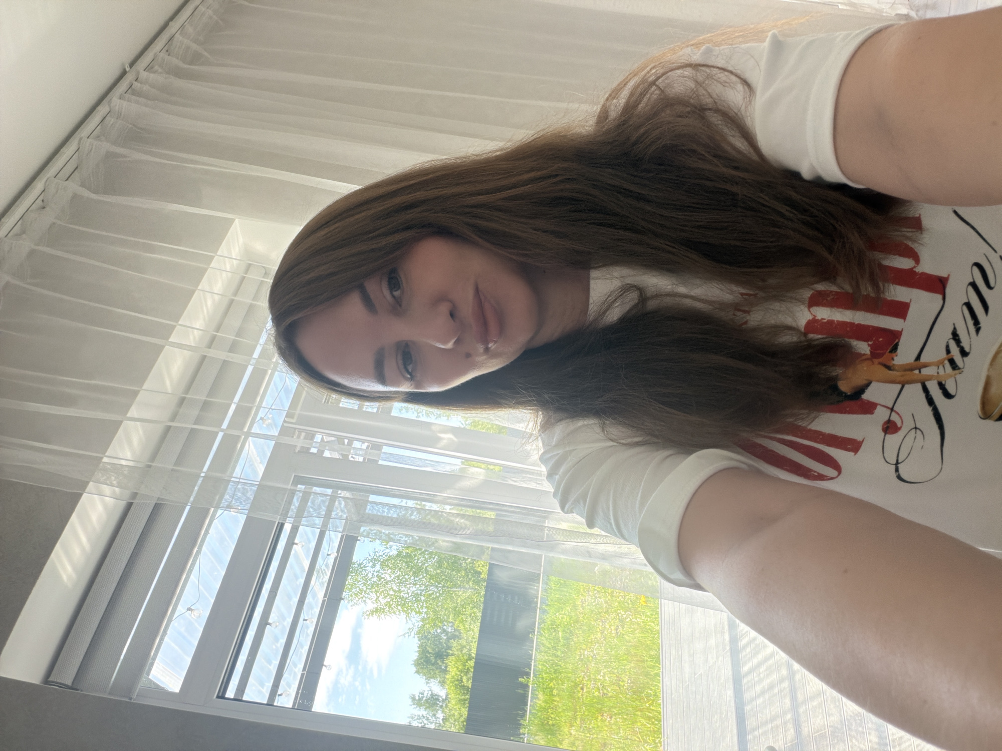 In Malinovka, Russia, a woman with long brown hair is taking a selfie indoors. She is wearing a white t-shirt with red lettering and an image of a deer. The background features a large window with sheer white curtains, revealing a glimpse of lush green trees and a bright blue sky outside. The sunlight streams through the window, casting shadows on the floor and the woman's arm. The room appears to be a residential space, possibly a living room or bedroom.