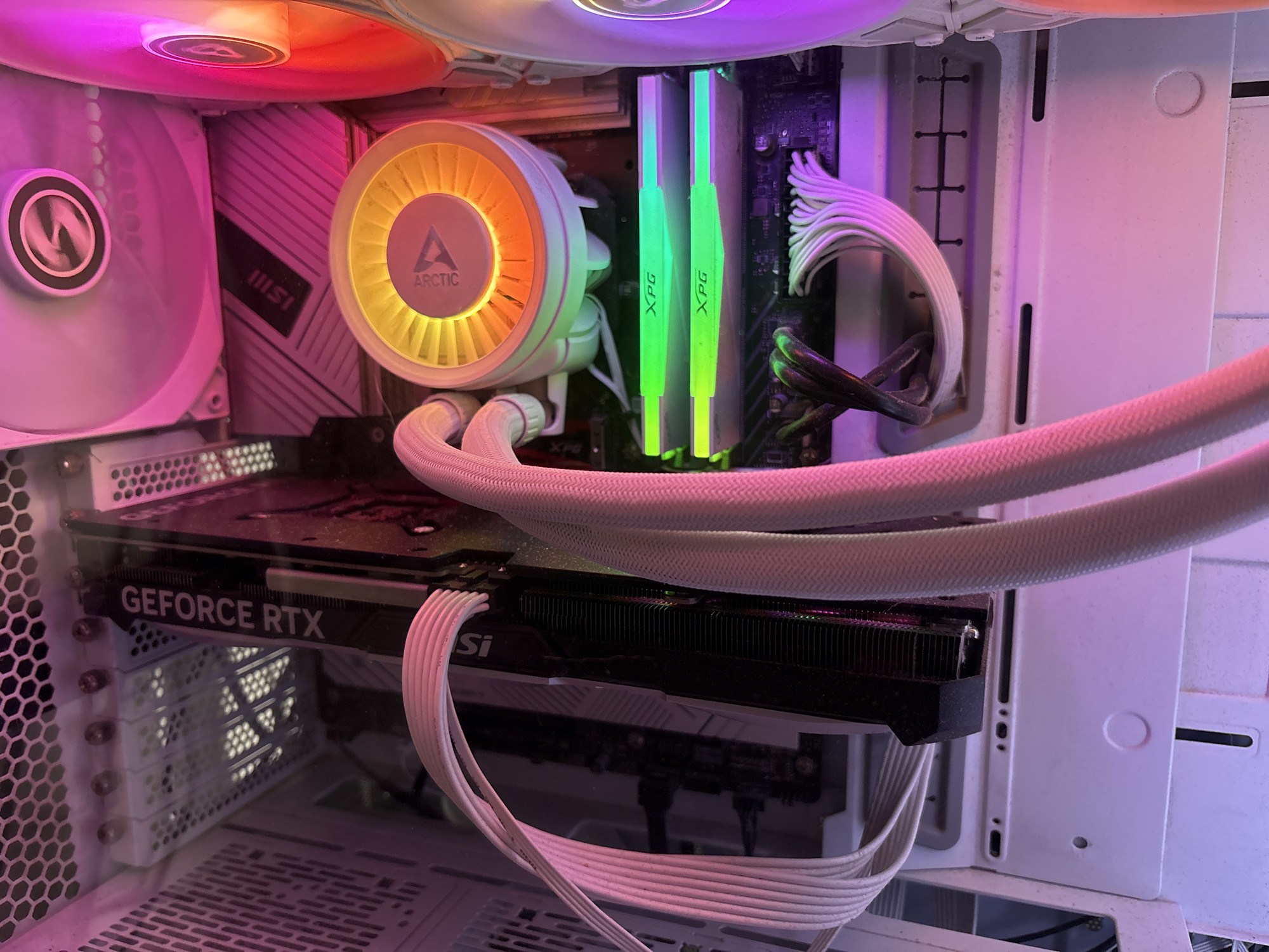 The image shows the interior of a white computer tower with RGB lighting. A black GeForce RTX graphics card is visible, along with two sticks of XPG RAM that are illuminated in green. A white CPU cooler with an orange and yellow illuminated fan is also present. White braided cables are neatly managed throughout the case. The lighting in the case is primarily pink and purple. There is no visible information within the image that would indicate its location in the city of Придорожный, Russia.