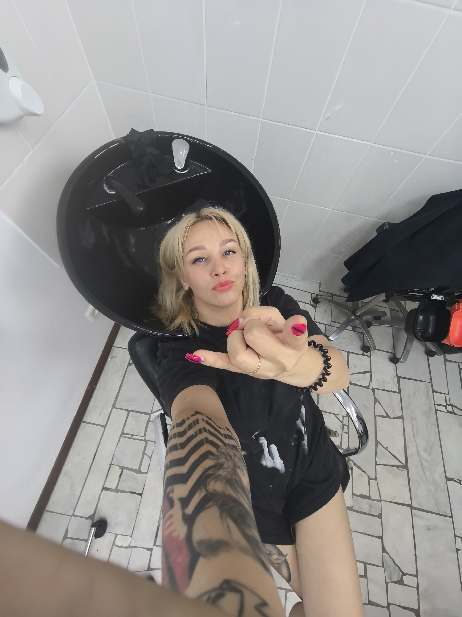 A young Caucasian woman with blonde hair and striking blue eyes is positioned in a hair salon's shampoo area, seated in a chair that faces away from the camera. She is captured in a selfie-style shot, with her arm extended towards the viewer. Her lips are painted with bright pink lipstick, and her fingernails are adorned with a matching vibrant pink polish. She wears a black t-shirt and black shorts. Her left arm, which is closest to the camera, displays a prominent tattoo. She is wearing a black beaded bracelet on her right wrist and a thin gold ring on her right ring finger. Behind her, a black shampoo sink is visible, and further back, a black drape covers part of a salon chair. The flooring is tiled with a geometric pattern of light and dark grey and white squares and rectangles. The walls are white and tiled. The scene appears to be a typical interior of a hair salon. The image does not provide any specific visual cues to pinpoint its exact location within the city of Chaikovsky, Russia.
