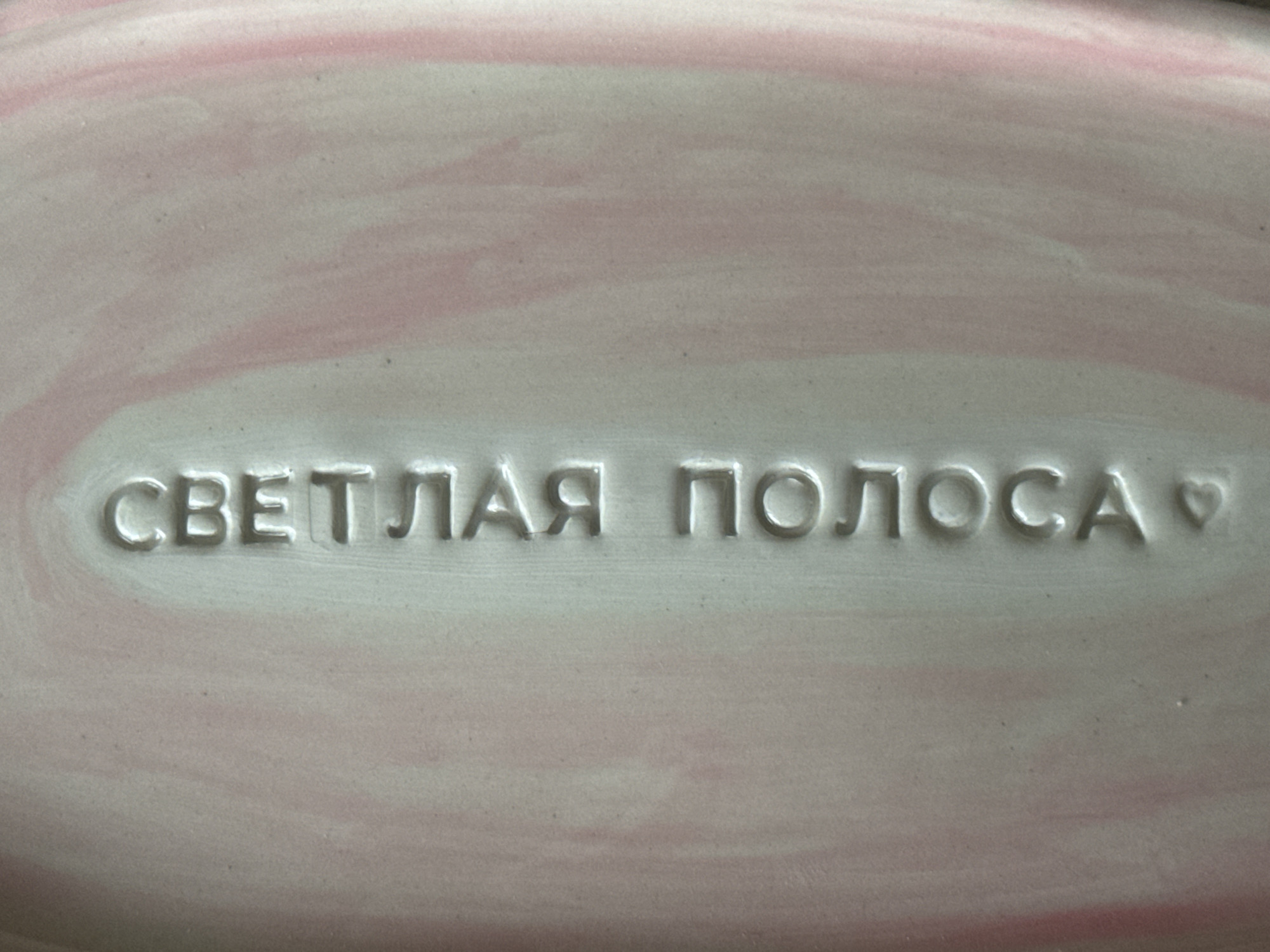 The image shows a close-up of a light pink surface with the Russian phrase "СВЕТЛАЯ ПОЛОСА" (SVETLAYA POLOSA) embossed in the center. The phrase translates to "Bright Streak" or "Bright Stripe". There's a small heart symbol embossed to the right of the phrase. The surface appears to be made of ceramic or similar material, possibly part of a plate or dish. The context of Saint Petersburg, Russia, is not evident from the image itself.