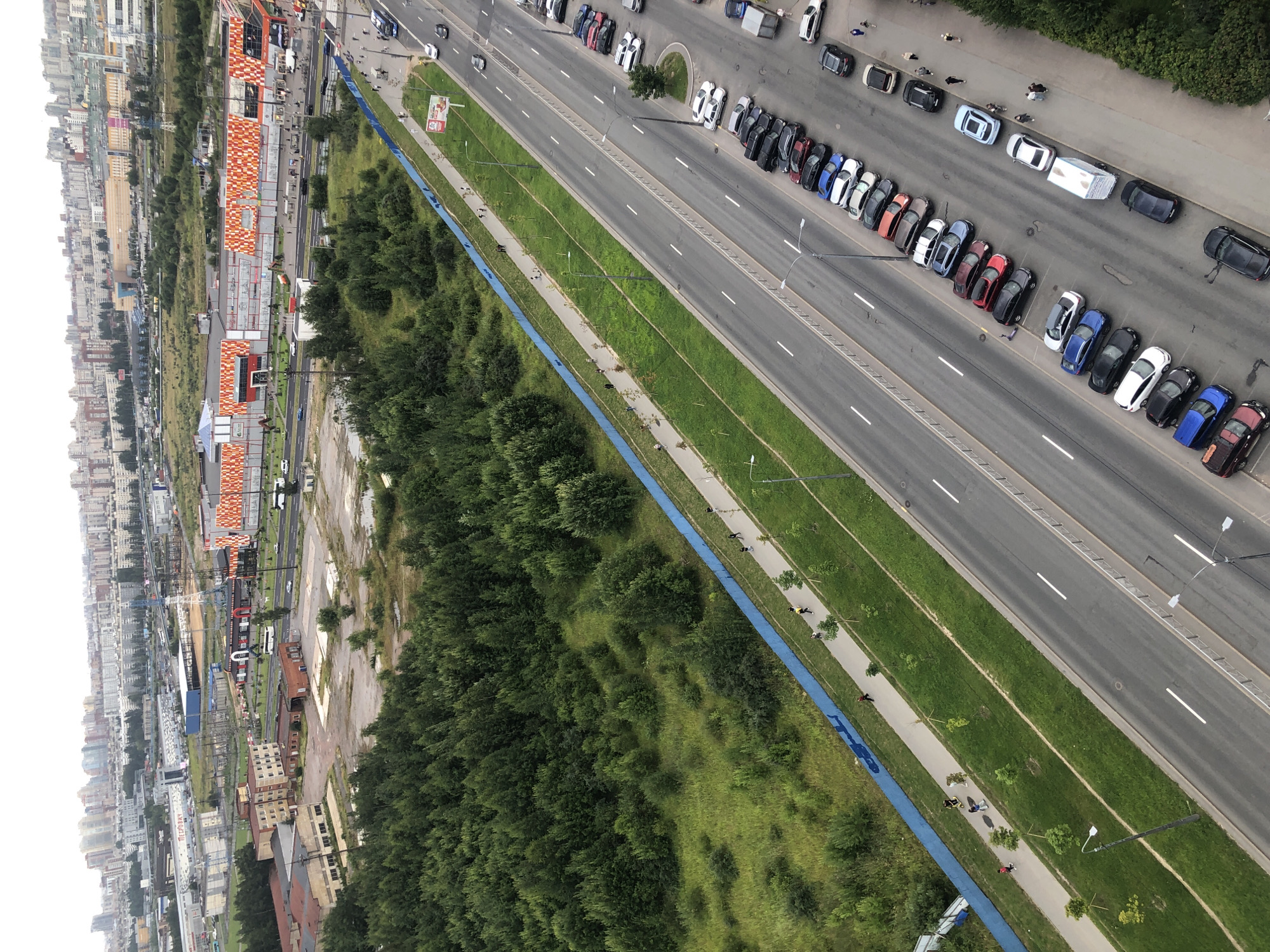 This is an aerial, high-angle, full shot of a city scene on a cloudy day. The foreground features a multi-lane asphalt road with parked cars on the right and a pedestrian walkway with people on the left, separated by a grassy median. The road runs diagonally from the bottom right to the top left of the frame. On the left side of the walkway is a dense green forest, bordered by a bright blue fence. Beyond the forest and road, there are various buildings, including a large shopping center with an orange and white patterned facade, industrial buildings, and residential high-rises in the distance. There are multiple cars parked along the road, and several people are visible walking or riding on the walkway, some of whom are on scooters. A few vehicles are driving on the road. The overall environment appears to be a mix of urban development and natural green spaces.