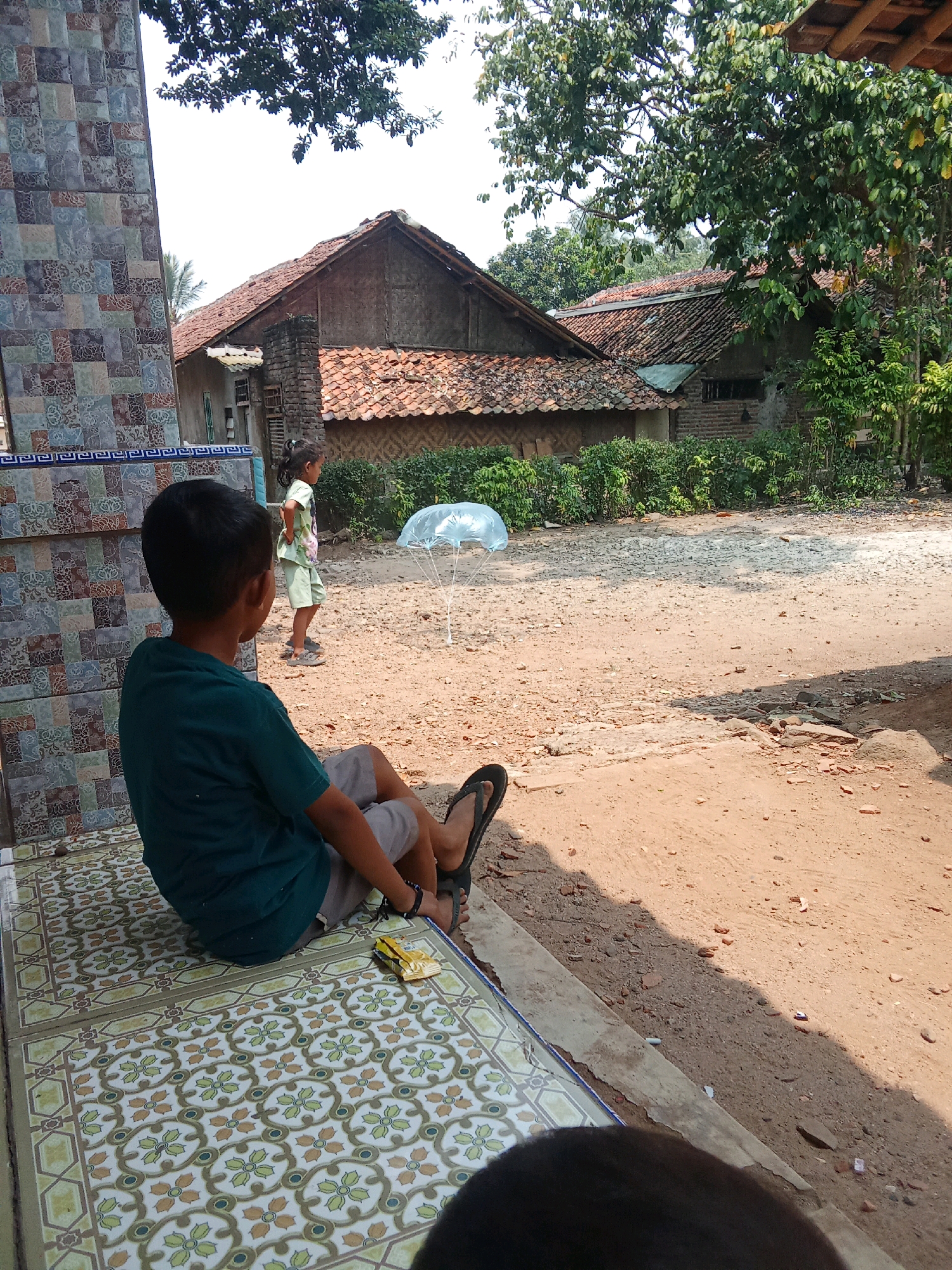In Cempaka, Indonesia, a young boy sits on a tiled porch facing away from the camera, wearing a green t-shirt and shorts. Beside him on the porch is a small yellow toy. In the background, a younger girl in a light green outfit stands on a dirt path, looking towards a clear plastic parachute suspended by strings. Traditional Indonesian houses with tile roofs and brickwork are visible behind the children, surrounded by lush green vegetation. The scene is illuminated by bright sunlight, casting shadows across the ground.