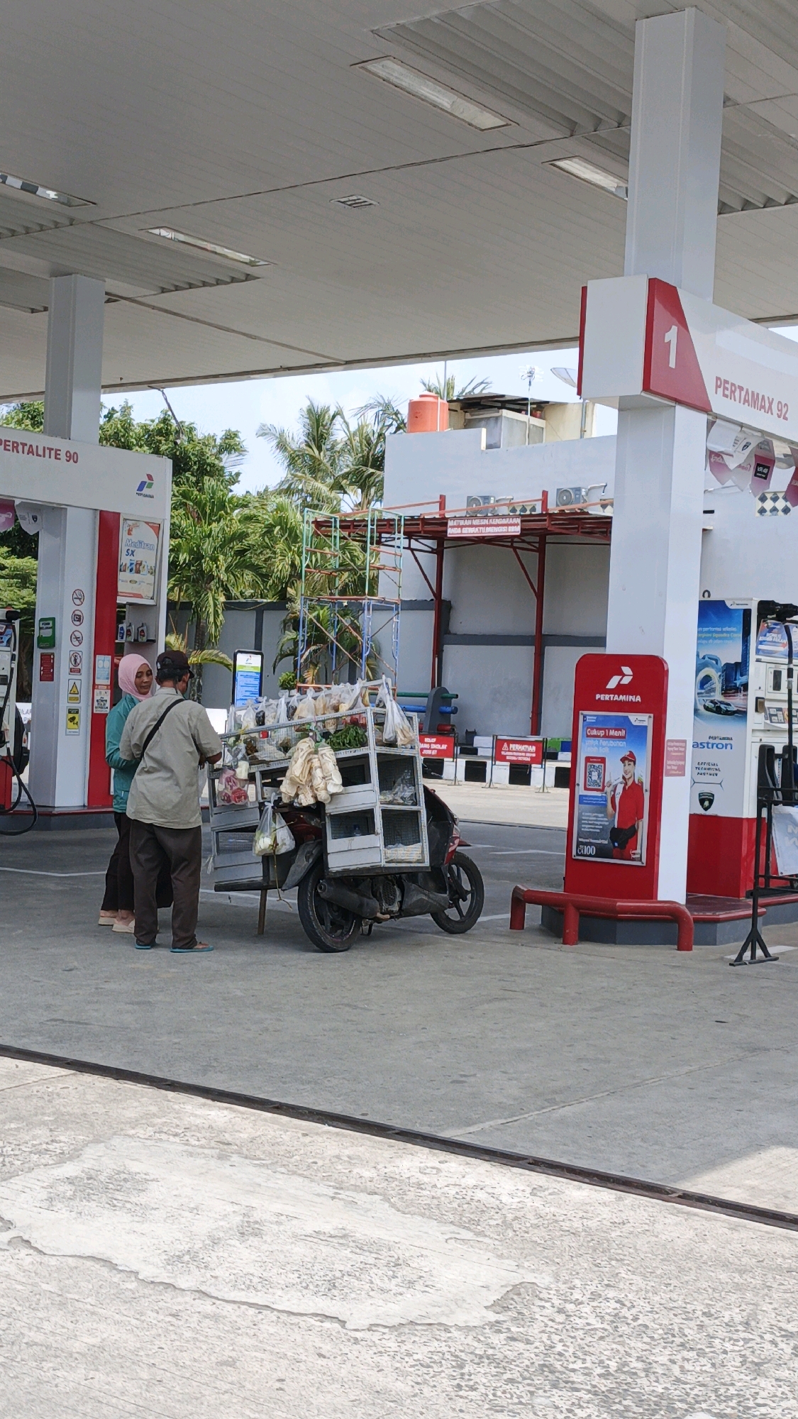 The image shows a street scene at a Pertamina gas station in Pangandaran, Indonesia. On the left, a white gas pump with "PERTAMALITE 90" signage is visible. In the center, a man wearing a light brown shirt and dark pants stands next to a motorcycle modified with a rack carrying various goods, including bags of groceries and produce. He is interacting with a woman wearing a teal long-sleeved shirt and a pink headscarf, who is standing to his left. A second woman in dark pants is partially visible to the left of the first woman. To the right, another Pertamina gas pump, labeled "PERTAMAX 92," is prominent, with red and white branding. A red bench and a display stand with advertising are also present. The environment consists of a paved ground surface, a covered canopy overhead supported by white pillars, and palm trees and buildings in the background under a clear sky. The overall scene captures a moment of commerce at a roadside service station.