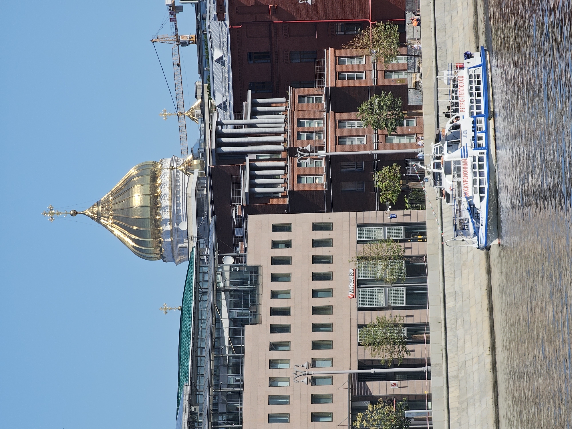 A wide shot captures a riverfront scene with a large, ornate golden dome of a cathedral dominating the skyline. To the left of the dome, a modern glass and concrete building with multiple floors and rectangular windows is visible. Adjacent to it, a red brick building with a unique architectural design featuring a colonnade of pillars rises. Further to the right, another red brick building with smaller windows is present. In the foreground, a white passenger boat with blue trim is cruising on the water. The boat has text in Cyrillic script on its side, indicating it is an excursion boat. The riverbank is lined with a stone embankment. The sky is clear and blue. Construction cranes are visible in the background behind the buildings.