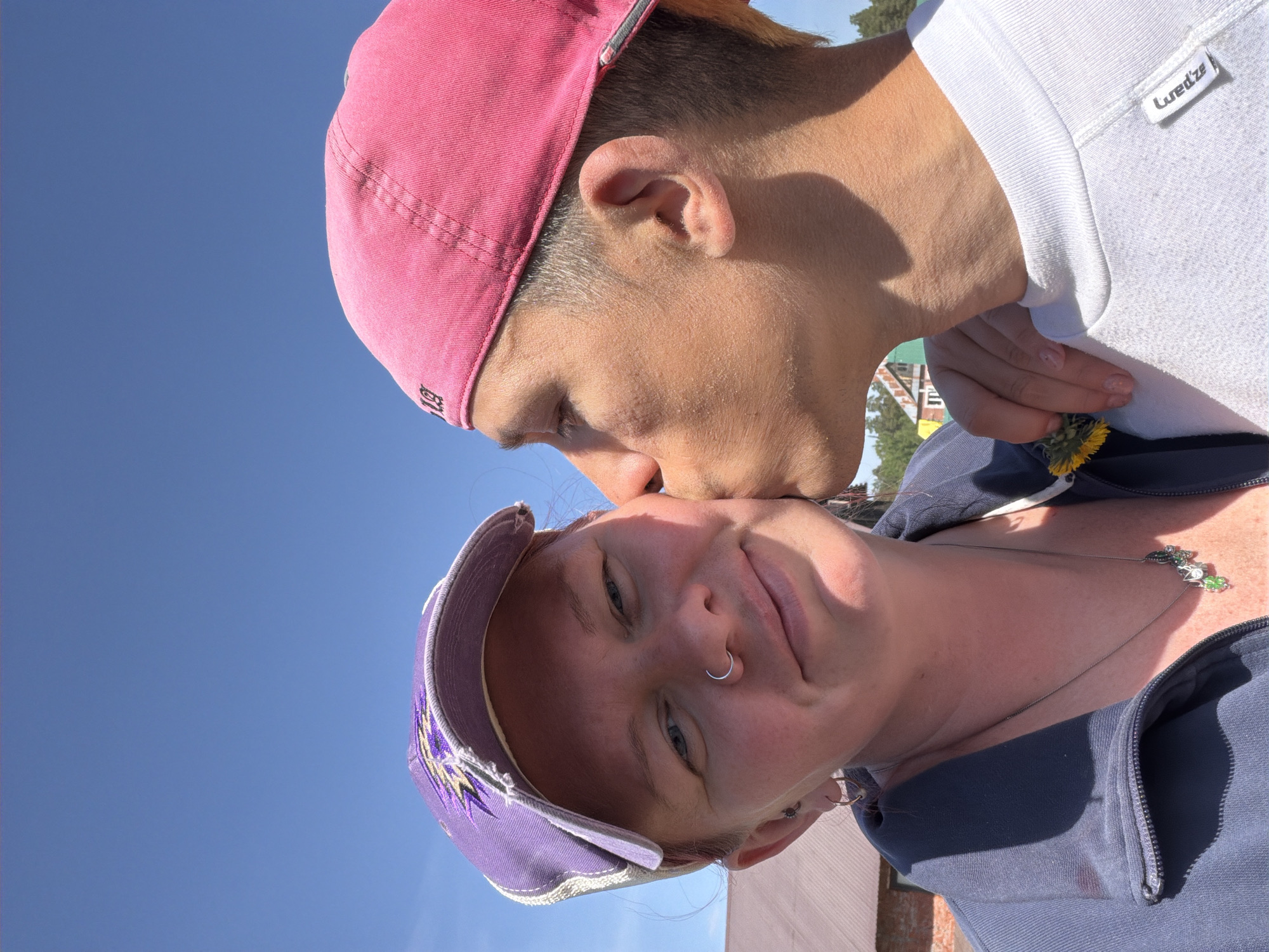 Two people are in the shot under a bright sky. The person in the foreground wears a purple baseball cap with white trim and designs, a nose ring, and a blue zip-up sweatshirt. They are looking at the camera and appear to be smiling. The other person is kissing the foreground person on the forehead. This person wears a pink baseball cap and a white sweatshirt with a "Luredze" brand label. They are holding a yellow dandelion flower between them. The location appears to be outdoors. While the image's exact location in Chaikovsky, Russia, cannot be confirmed, the background includes elements consistent with an outdoor environment in a park or recreational area.
