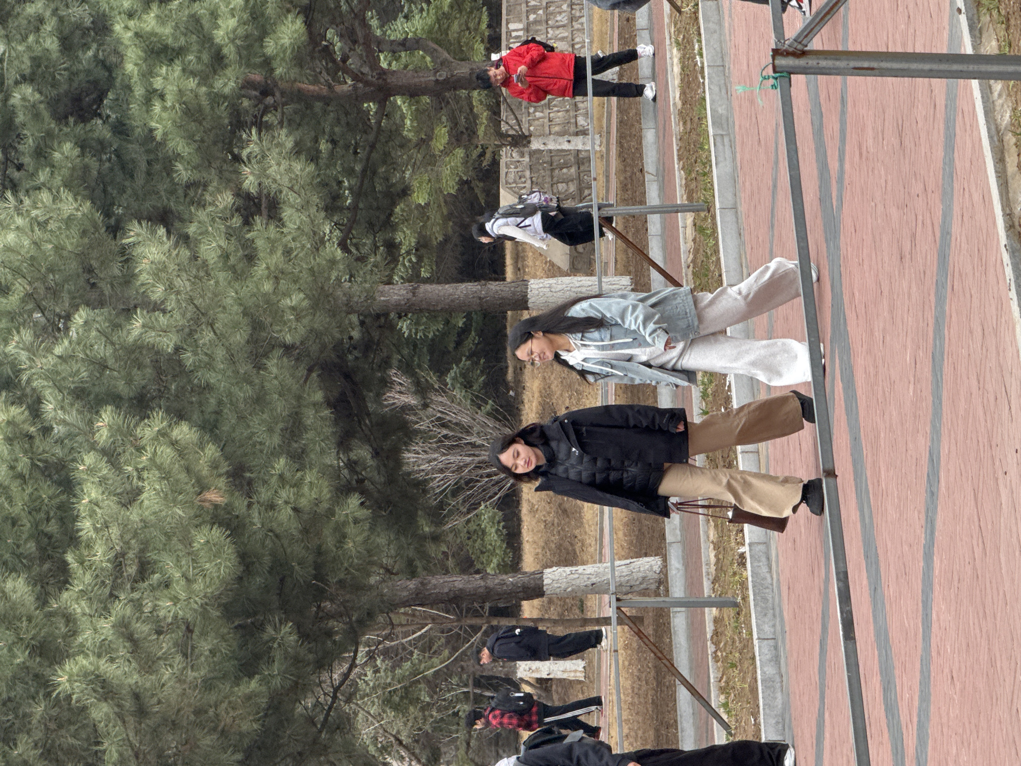 The image shows a group of people walking on a brick-paved walkway in an outdoor area, likely a park. There are trees to the left, with dense green foliage. A metal railing runs along the walkway. Several individuals are visible, including two women in the foreground. One is wearing a dark jacket and beige pants, carrying a purse. The other woman wears a light denim jacket and light-colored pants. Other people are further back, some wearing jackets. In the background, a person in a red jacket is visible. The location appears to be an urban park setting. The specific city or location within China is unknown.