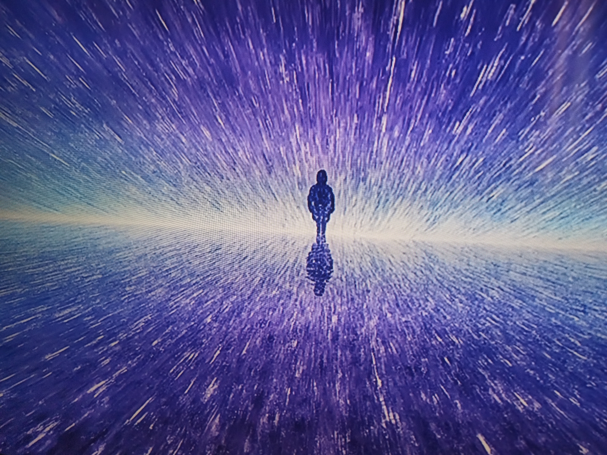 A lone figure stands with a reflection on a surface of purplish-blue with radiating white streaks. The figure's dark silhouette contrasts against the bright horizon line, creating a sense of depth. The streaks give the impression of movement or energy emanating from the center of the image. There is no indication in the image that this photograph was taken in Kramatorsk, Ukraine.