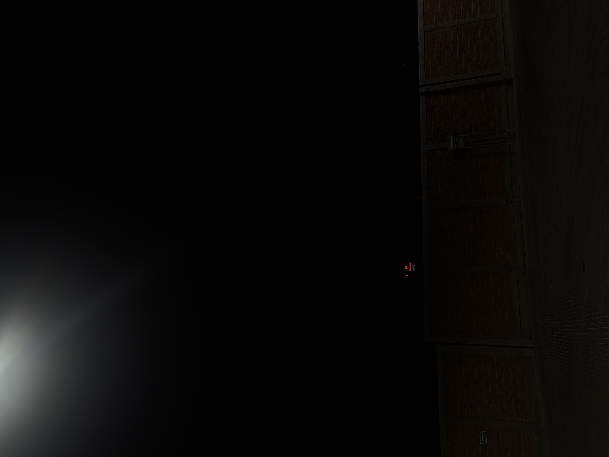 The image appears to be taken at night or in very low light conditions, likely indoors based on the presence of what seems to be a textured wall or stack of large, potentially brick-like objects framed by a dark metal or similar structure. This structure extends vertically, divided into sections. There might be a small arrow or indicator visible near the bottom. The scene is poorly lit except for a bright light source near the bottom-left of the frame, creating a very high contrast image. The details are largely obscured by darkness. There is no explicit information present to confirm location.