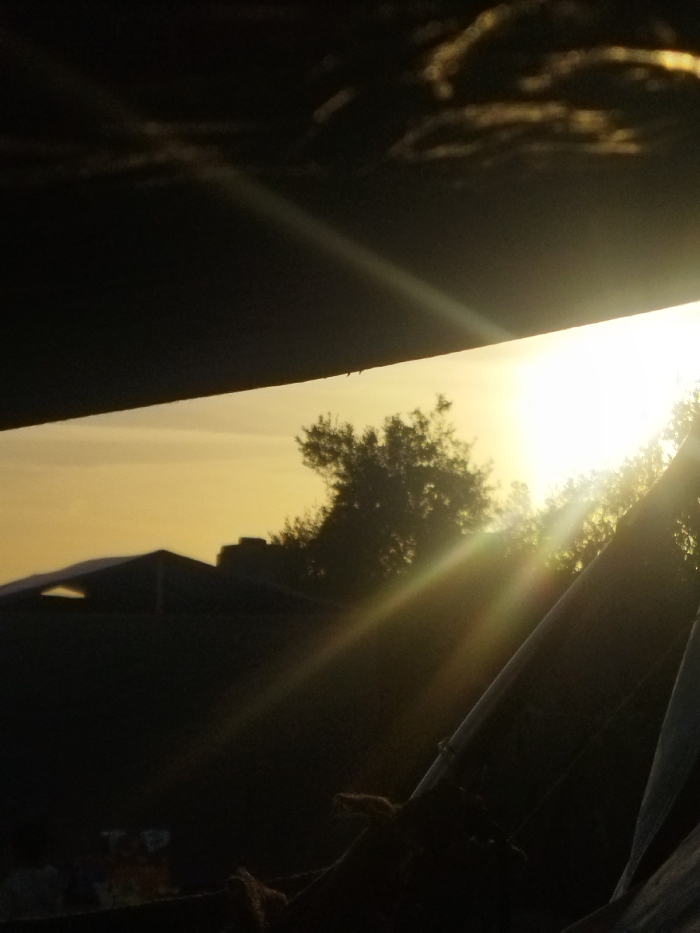 The image displays an outdoor scene in Khan Yunis, Palestinian Territory, at either sunrise or sunset. A strong, low-angle light source, presumed to be the sun, is positioned on the right side of the frame, emitting intense white and yellow light that generates prominent diagonal lens flares across the foreground and midground. The sky exhibits a gradient of yellow-orange hues, fading into darker tones towards the upper left where indistinct cloud formations are visible.
A dark, broad, horizontal structural element occupies the top portion of the frame, angled slightly downwards from left to right. Below this, silhouetted against the bright sky, are several structures: a building with a distinctive pitched roof and an opening or feature on its left side, and a smaller, rectangular structure positioned to its right. A large tree with dense, dark foliage is silhouetted in the midground, positioned to the left of the sun's direct light. The lower right foreground reveals parts of an informal structure, possibly a tent or shelter, consisting of dark fabric or tarp secured with visible ropes around diagonal poles. Indistinct bundles or objects are faintly discernible in the dark lower left foreground. No human figures or discernible actions are present in the scene.