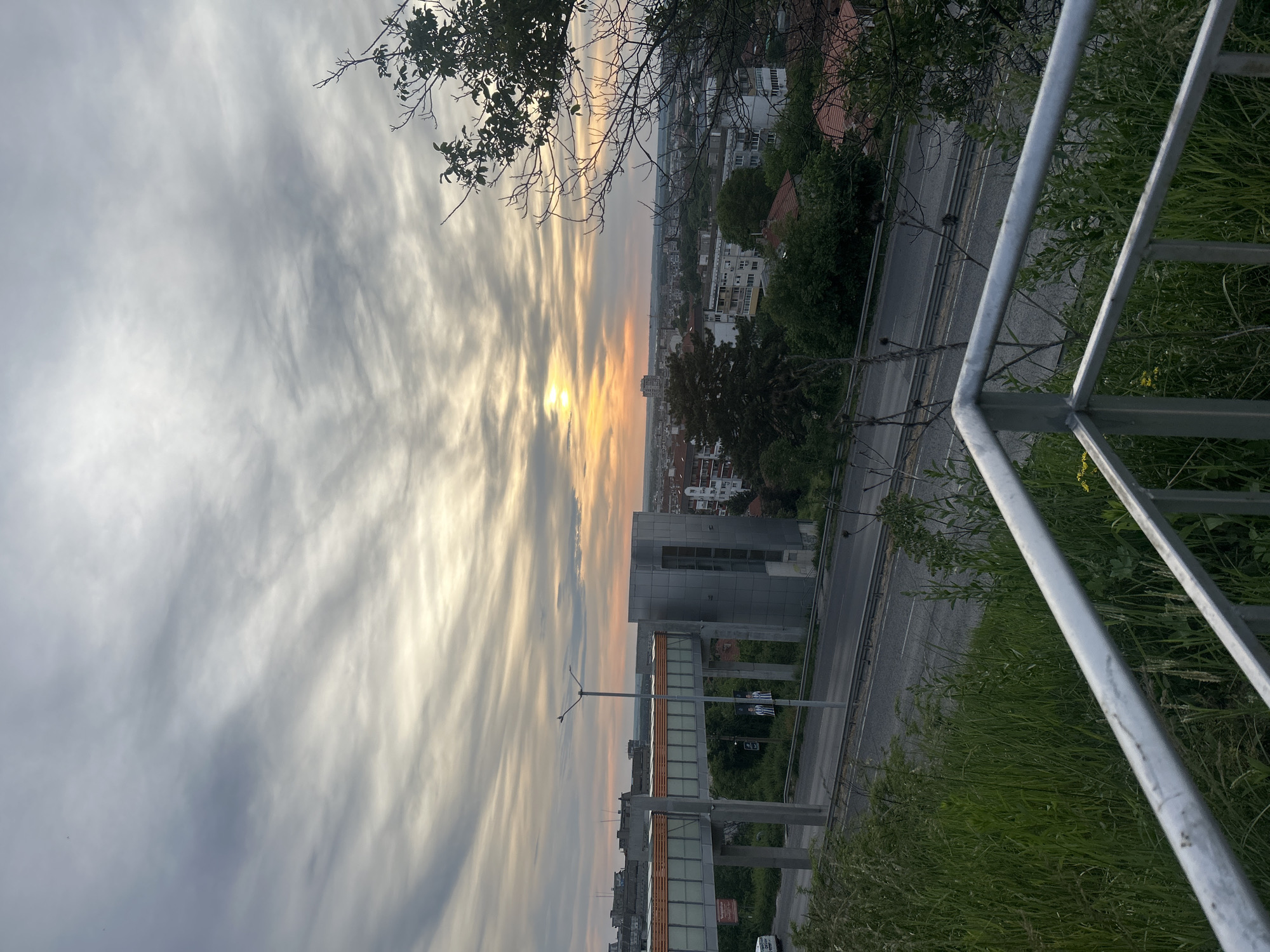 The image shows an urban view at sunset in Ruse, Bulgaria. A pedestrian overpass spans a road. Buildings, trees, and a metal railing are visible. The sky is filled with streaky clouds and the sun is low on the horizon.