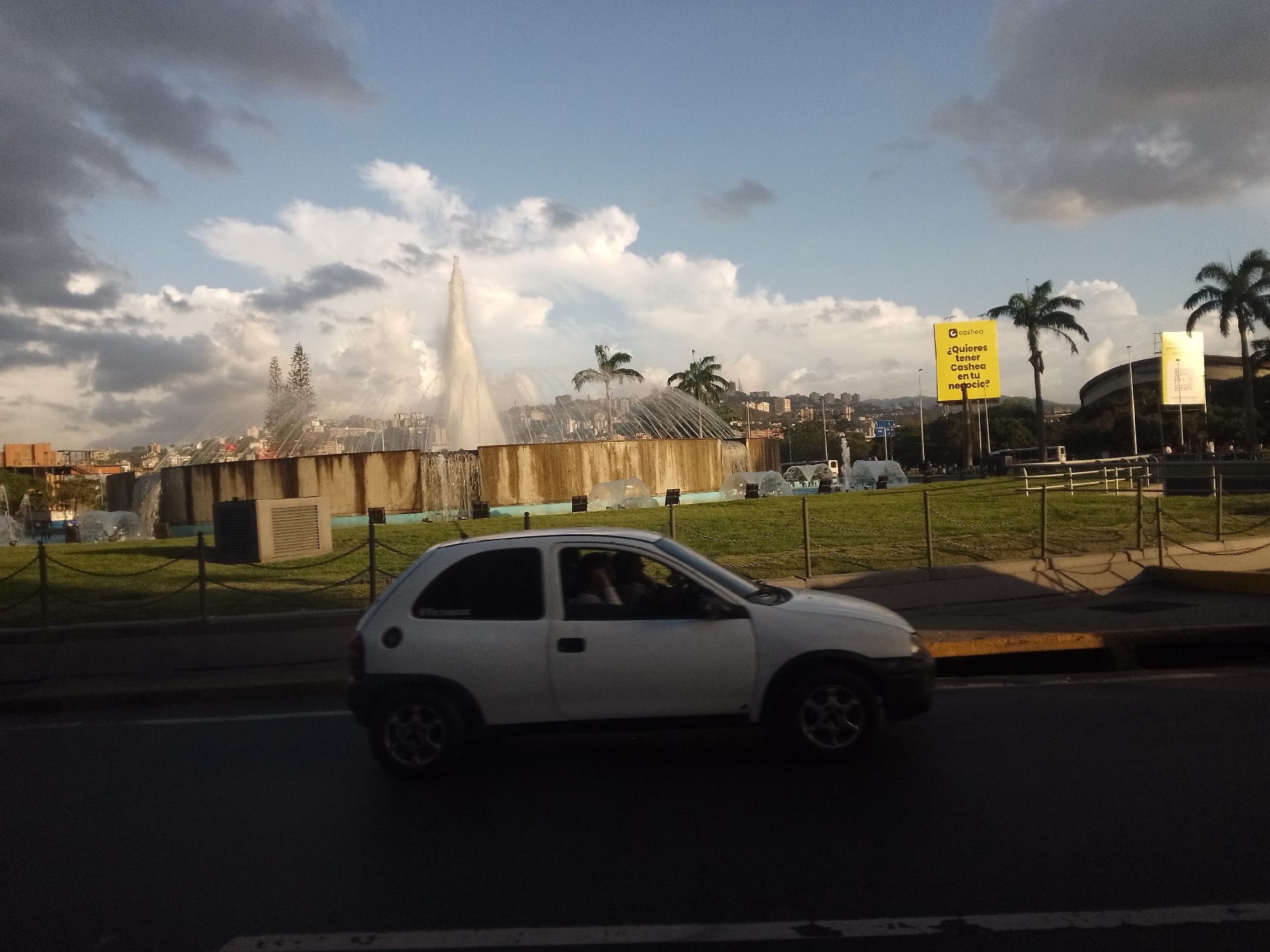 This media file depicts an urban street scene featuring a white two-door hatchback car driving on a dark asphalt road in the foreground. A person's arm is visible inside the car, likely the driver. The road is bordered by a curb and a grassy median, separated by a fence composed of posts and chains.
In the mid-ground, a prominent, multi-tiered fountain complex is actively spraying water high into the air. The fountain is set within a well-maintained green lawn, enclosed by a low concrete wall. Several palm trees are scattered around this area.
The background reveals a dense cityscape with numerous buildings stretching across hilly terrain, typical of a large urban area. The sky is partly cloudy, displaying patches of bright blue interspersed with darker, gray clouds. The lighting suggests late afternoon or early evening, with warm sunlight illuminating the upper parts of the scene, including the fountain spray and some clouds, while the foreground road and the lower part of the car are in shadow.
Two large billboards are visible in the background. The most distinct one is yellow and features the brand "cashea" with the Spanish text: "¿Quieres tener Cashea en tu negocio?" (Do you want to have Cashea in your business?). The overall scene portrays a dynamic urban environment with a blend of natural and architectural elements.