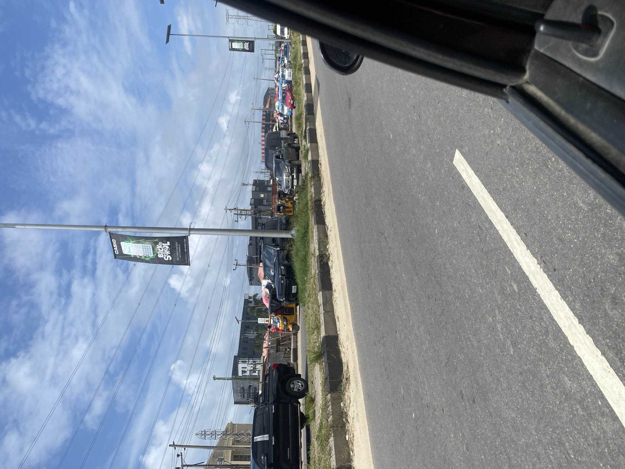 The image depicts a street scene in Lekki Peninsula II, Nigeria, under a partially cloudy sky. In the foreground, the asphalt road with a white lane marker is visible, and a portion of a dark-colored car's interior, including the side mirror and door frame, is in the lower right corner.
Further down the road, several vehicles are present. A black Toyota Tundra pickup truck is parked on the left side of the road, beside a grassy verge with a concrete curb. To its right, a yellow auto-rickshaw is parked, with a person standing next to it. Beyond the auto-rickshaw, a dark-colored SUV and a silver pickup truck are visible.
In the background, a line of other vehicles, including cars and possibly minibuses, are observed. Several commercial buildings and structures, some appearing modern and multi-story, are situated along the street. Overhead, power lines crisscross the sky, and a tall lamppost with a banner advertising "Casio Back to School" is prominently displayed on a pole. The overall impression is of a busy urban environment.