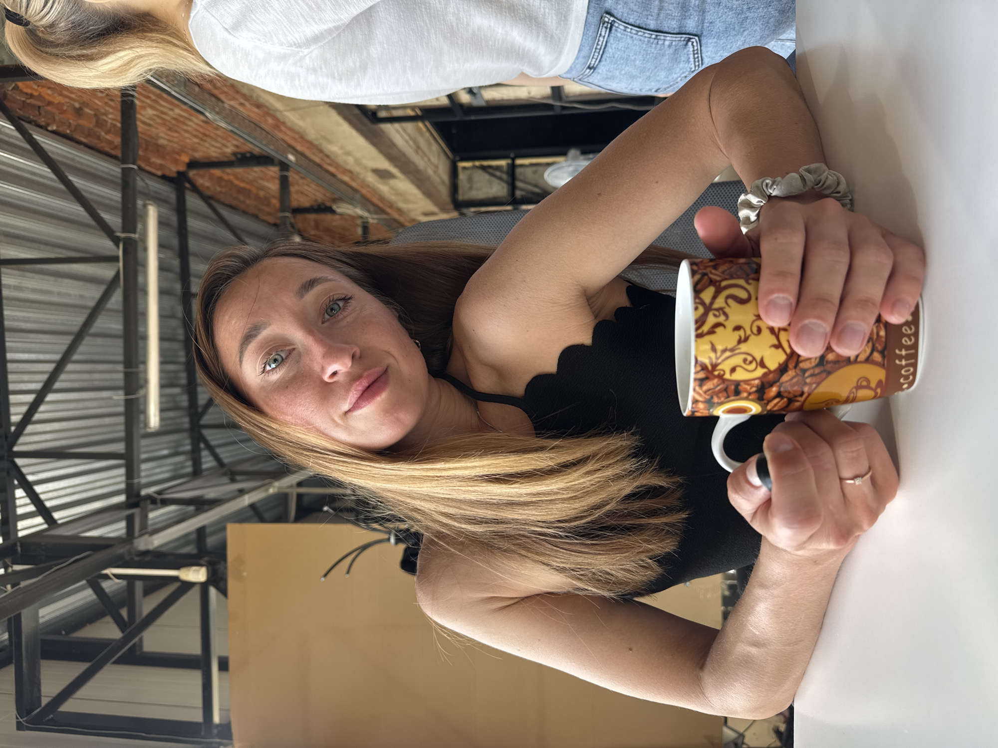 A woman with long blonde-highlighted hair is sitting at a white table, holding a coffee mug with a design of coffee beans and the word "coffee" printed on it. She is wearing a black top with a scalloped neckline. She is looking at the camera. She has light eyes and fair skin. A beige scrunchie is on her wrist. A blonde-haired woman is partially visible in the upper left corner, wearing a white shirt and blue jeans. The background shows industrial-style ceilings with metal beams and exposed brick. The location may be the city of Chaikovsky, Russia.