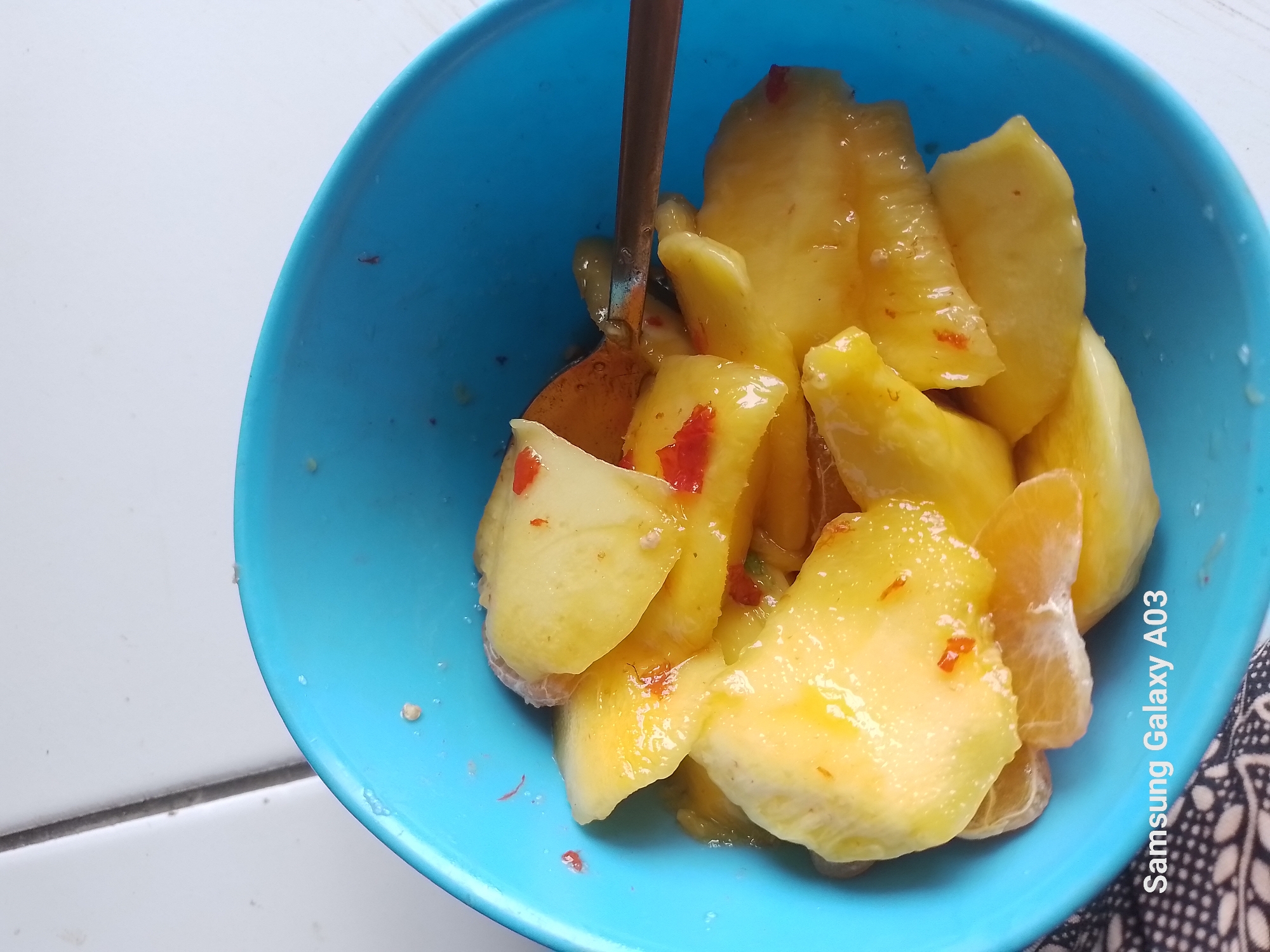 A close-up, overhead shot displays a bright blue bowl filled with sliced fruits, possibly mangoes and oranges, coated in a glistening sauce. Small, red pieces, likely chili flakes, are mixed throughout the fruit, suggesting a spicy preparation. A wooden spoon rests within the bowl, partially submerged in the fruit. The bowl is positioned on a white surface with a thin, dark line running vertically down the left side, which appears to be a seam or grout line of a tiled surface. The lower left corner of the image features the text "Samsung Galaxy A03". The context of Kali Asin, Indonesia, suggests this might be a dish prepared or consumed in that region, though no specific landmarks are visible.