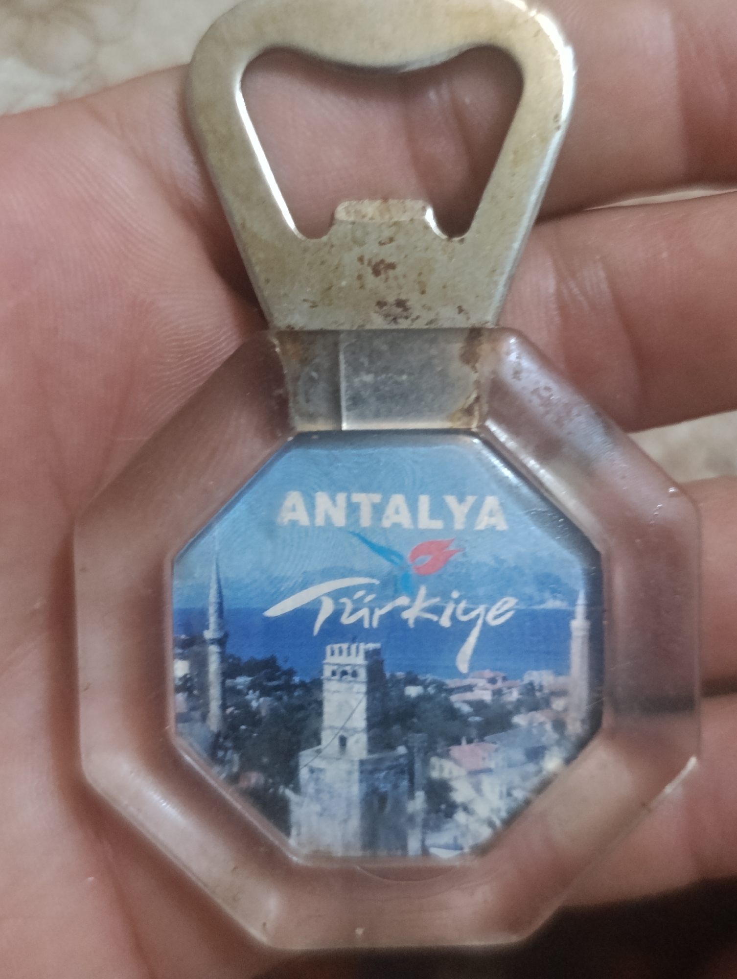 The image shows a close-up of a hand holding a bottle opener keychain. The keychain features a clear plastic octagonal frame with a printed image of a city skyline. Text on the image reads "ANTALYA" and "Türkiye" in white lettering. The skyline depicts buildings and minarets against a blue sky, suggesting a Turkish city. The bottle opener is made of metal, appearing aged and slightly tarnished. The background is blurred, showing skin texture and hints of a light-colored surface. There is no indication of the city of Baku, Azerbaijan in the image.