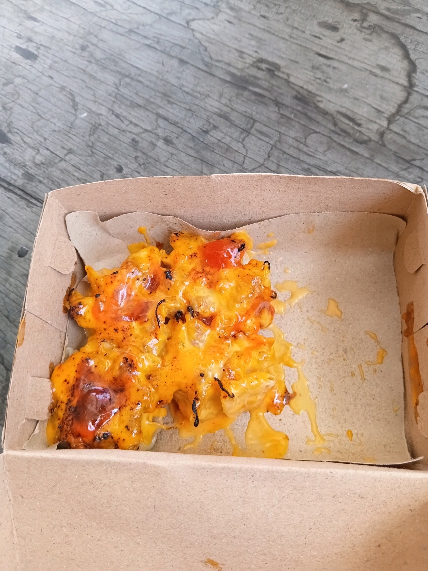 The image shows a top-down view of a cardboard box containing a portion of macaroni and cheese. The macaroni and cheese is topped with melted orange cheese and appears to have been recently served. The box is open and sits on a grey, weathered wooden surface. There are no people visible in the image. The scene appears to be outdoors, as indicated by the natural lighting and the texture of the wooden surface, which could be part of a table or bench. The location of Batu Belaman, Indonesia is not explicitly depicted in the visual elements of the image but is provided as contextual information.