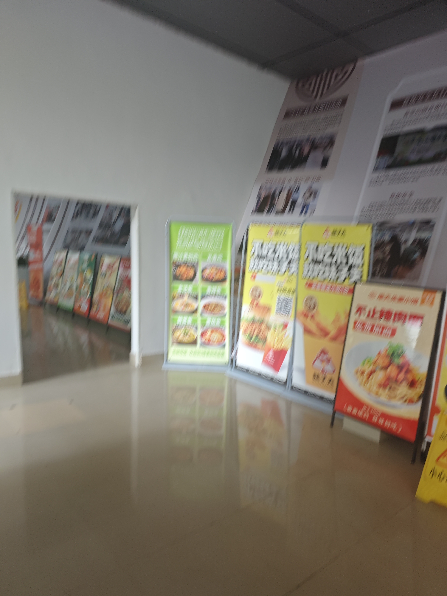 The image depicts an indoor scene, likely a public space with advertisements displayed on vertical stands. The setting appears to be a corridor or hallway with a polished, reflective floor. On the right side of the image, there are several large, colorful display boards. One of these, on the far right, is yellow with a red symbol and some text. Adjacent to it is a red and white sign featuring an image of a pasta dish. To the left of these, there are two yellow signs with images of food items and QR codes. Further left, a green sign displays multiple images of various dishes. In the background, visible through an opening on the left, is a row of more display boards, some with orange and green elements. The overall impression is that of a commercial area, possibly a food court or a marketplace. The lighting is somewhat dim, and the image is slightly blurred, suggesting it was taken in motion or in low light conditions. There are no identifiable people clearly visible in the scene. The environment suggests a modern, possibly commercial building.