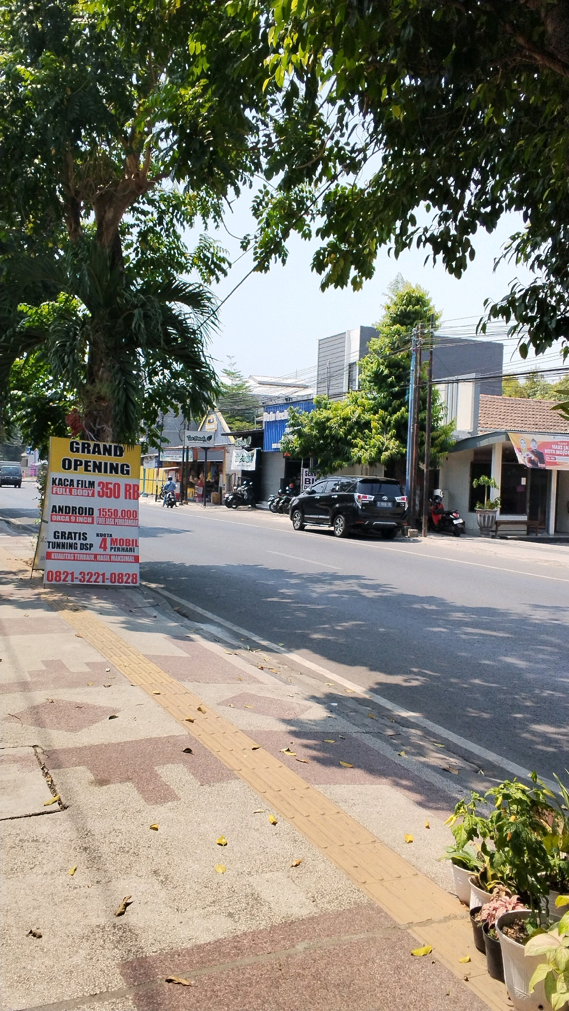 A bright, sunny daytime street scene is depicted in Banjaragung, Indonesia. The image is dominated by a tall, yellow A-frame sign on the left side of the sidewalk, advertising a "GRAND OPENING" with various product and service offers in Indonesian. The sign lists prices for "KACA FILM FULL BODY" (full body window film) and "ANDROID" devices, along with details about "GRATIS KUOTA" (free data quota) and "4G MOBIL" (4G mobile). A phone number, "0821-3221-0828", is prominently displayed.
The sidewalk in the foreground is paved with textured concrete and features a tactile paving strip for the visually impaired. Fallen yellow leaves are scattered across the sidewalk and road. Beyond the sidewalk, a road runs horizontally across the image, with a black SUV parked on the right side. Several motorcycles are visible parked in front of shops lining the street.
The background shows a commercial street with various buildings, some appearing to be shops or businesses. Trees with lush green foliage line the street, providing shade in some areas. Electrical wires are visible strung between poles. The sky is clear and bright blue. The overall impression is a typical urban commercial area on a sunny day.