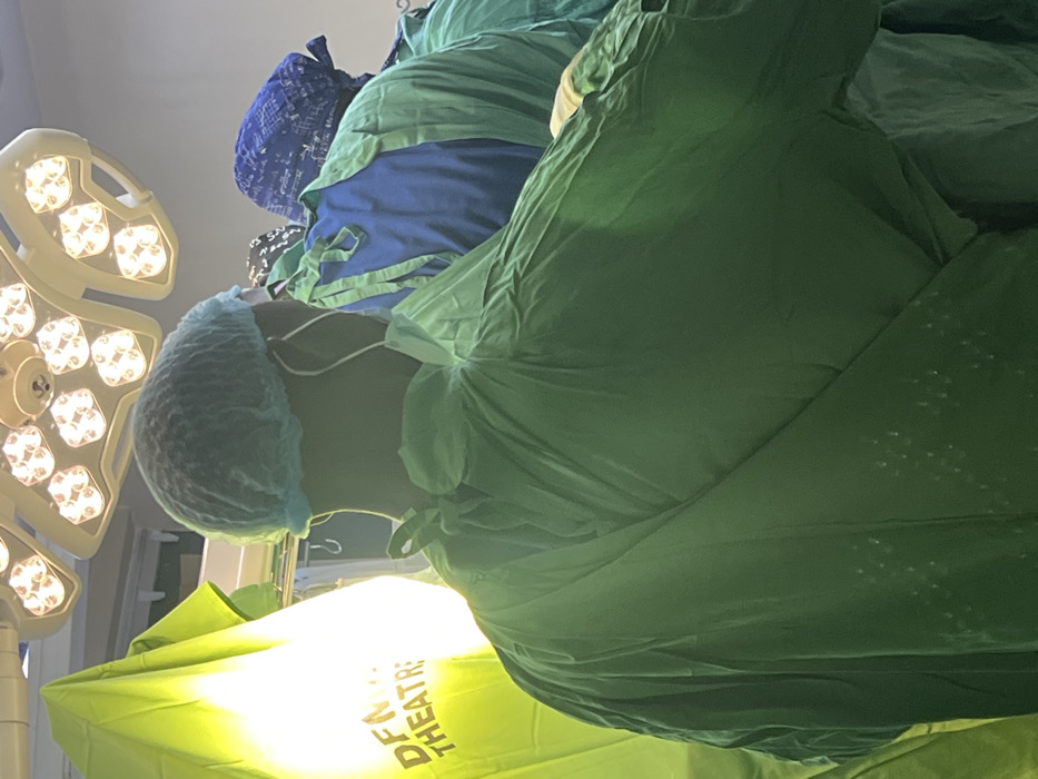 The image depicts an operating theater at DFM Hospital in Nairobi, Kenya. In the foreground, a medical professional is seen from the back, wearing a light blue bouffant cap, a medical face mask with straps visible around the ear, and a dark green surgical gown tied at the neck. To the right and slightly behind, another individual wears a blue patterned head covering and a green surgical gown over a blue scrub top. A third person in a green gown is partially visible further to the right. Above, a multi-panel overhead surgical light fixture illuminates the scene. On the left, a bright yellow-green medical drape or screen is partially visible, bearing the text "DFM" and "THEATRE" in dark letters. The individuals appear to be positioned for or engaged in a medical procedure.