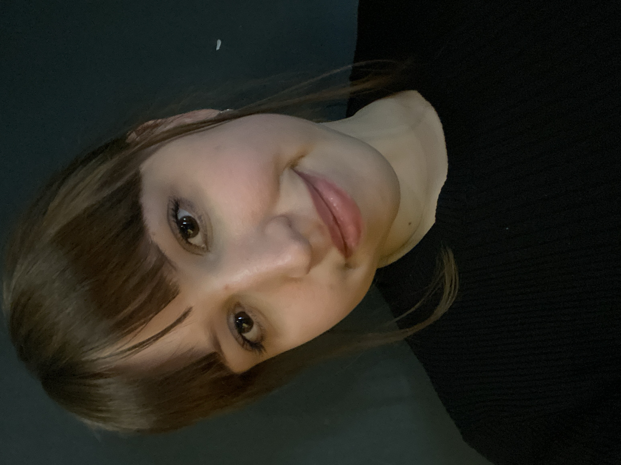 A close-up portrait of a young woman with light skin and brown hair with bangs. She is wearing a black top. The background is dark and indistinct. The location is unknown, and there is no information to suggest that the photo was taken in Chaikovsky, Russia.