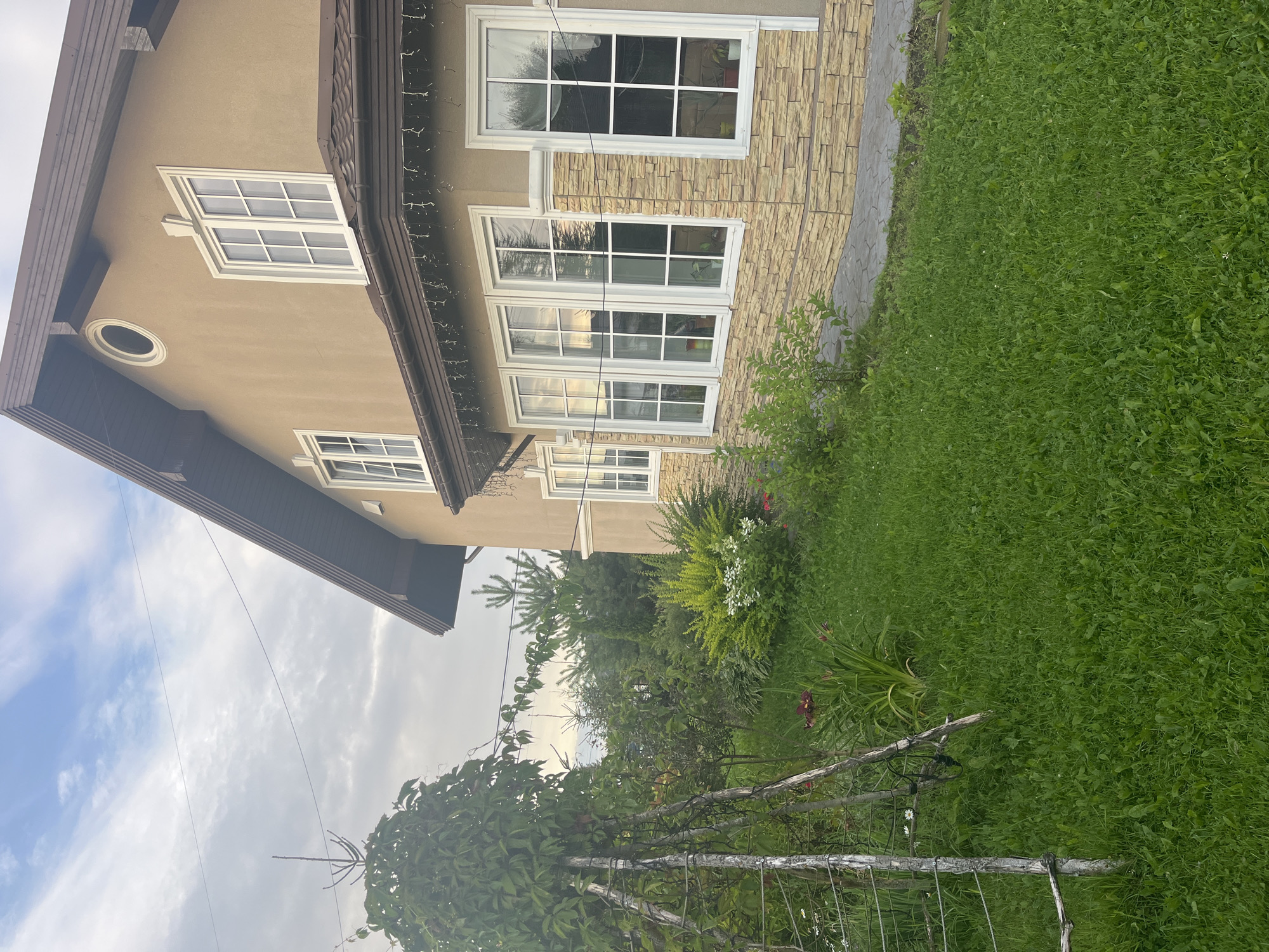 The image depicts the exterior of a two-story house in Vladimirovka, Russia. The house has a light brown stucco exterior with a dark brown roof and brown trim around the windows. The windows are white and divided into multiple panes. The lower portion of the house is accented with light brown stone siding. In front of the house is a well-maintained green lawn with several trees and bushes, including a trellised plant on the left side of the frame. A pathway made of irregular stone slabs leads to the house. The sky is cloudy, with patches of blue visible. The overall scene suggests a residential property in a suburban or rural setting. There are no people or animals visible in the image.