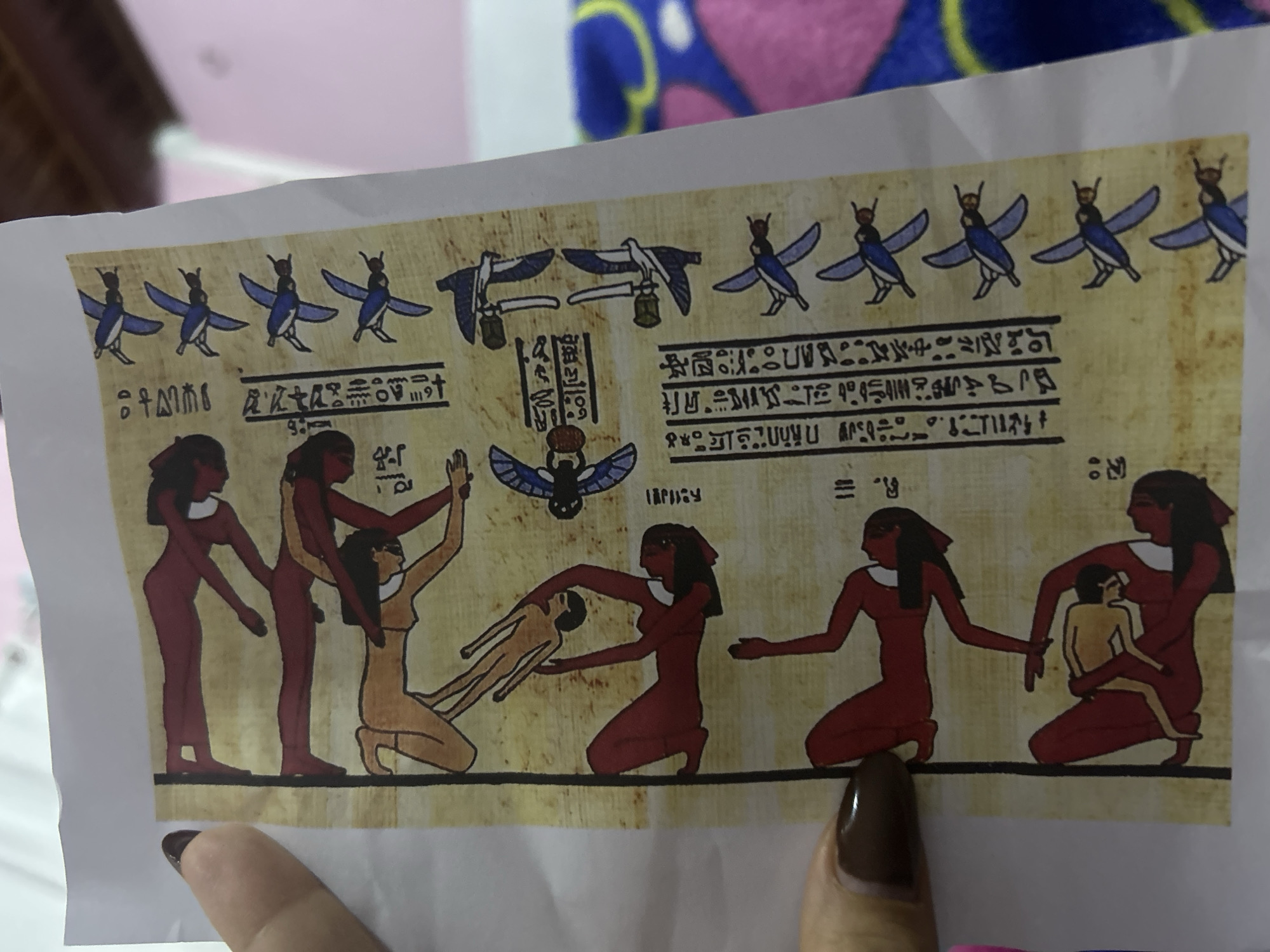 The image shows a reproduction of an Egyptian papyrus painting. The papyrus depicts a scene with several figures. On the left, three figures are shown, two in reddish-brown and one in a lighter tone. In the center, a reddish-brown figure is kneeling and appears to be holding or supporting the lighter-toned figure. To the right, two reddish-brown figures are shown, one holding a child. Hieroglyphic writing and stylized depictions of birds adorn the top of the image. The papyrus reproduction is being held by a person with manicured fingernails. The location is not evident from the image.