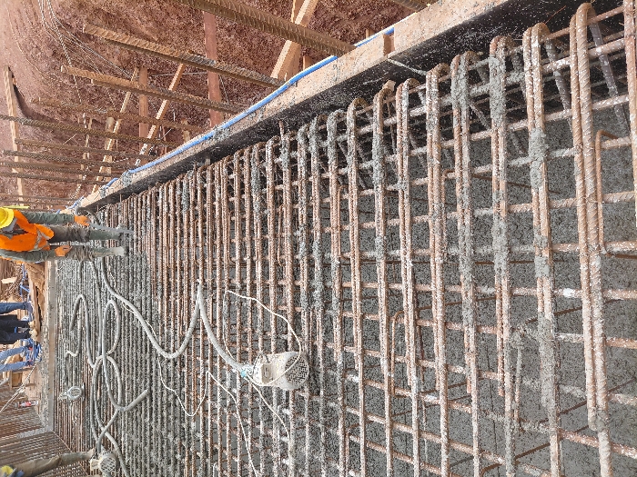 The image depicts an active construction site in Fdalate, Morocco, focusing on a concrete pouring operation. In the foreground and midground, a grid of steel rebar, serving as structural reinforcement, is partially covered with freshly poured, wet concrete. Several concrete vibrators, connected by hoses and cables, are laid across the rebar mesh, with one prominently visible in the lower-left.
In the mid-right, a construction worker is positioned, bending over. This individual wears a yellow hard hat, an orange safety vest over a green long-sleeved garment, work gloves, and dark boots. Their specific action is unclear, but they appear to be inspecting or working with the concrete or rebar. Another worker's lower legs and feet, clad in light-colored boots, are visible on the rebar in the far-left. In the background, several other individuals are partially visible, standing on what appears to be a higher, stable ground level or a completed section of the structure.
To the right of the concrete work, a deep excavation with reddish-brown earth walls is visible. Wooden planks and vertical rebar elements are arranged along the excavated wall, functioning as shoring or formwork. A blue pipe runs horizontally along the upper edge of this excavation.