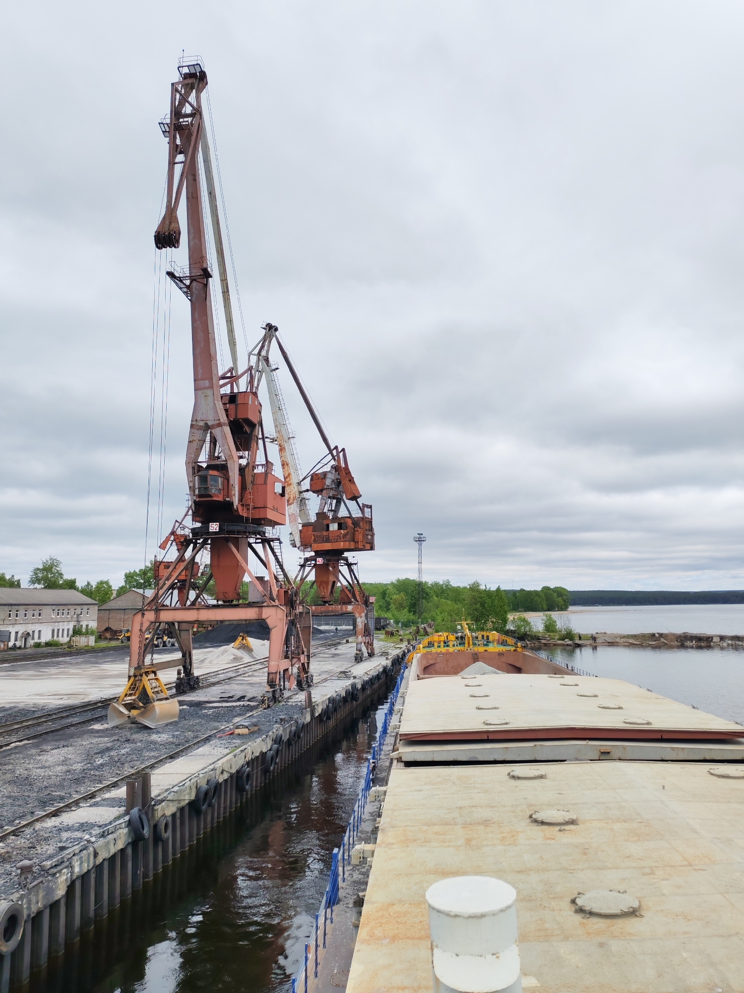 The image depicts an industrial port scene in Medvezhyegorsk, Russia, under an overcast sky, likely on Lake Onega. The central subjects are two large, aged, orange-red industrial gantry or portal cranes positioned along a concrete quay. The crane on the left features a substantial open clamshell grab bucket, and "52" is visible on its cabin. The crane on the right also displays "43" on its cabin. Both cranes show considerable rust and wear.
Piles of dark aggregate, resembling coal or gravel, are scattered across the dock, with a smaller, lighter pile of sand or gravel further back, where a yellow excavator is operating. The quay itself is concrete, reinforced with vertical pilings and worn rubber fenders, reflected in the dark water alongside.
A long, flat-decked barge or cargo ship is moored on the right, its deck covered with numerous round hatch covers. The yellow bow section of the vessel appears to be loaded with bulk material. In the background, a forested shoreline stretches along the lake under the cloudy sky, revealing a sandy beach area. Two small, distant figures are visible on the far shore. On the left, several old, multi-story industrial buildings with multiple windows complete the utilitarian setting. The scene suggests ongoing cargo handling operations in a well-established, though aged, industrial port.