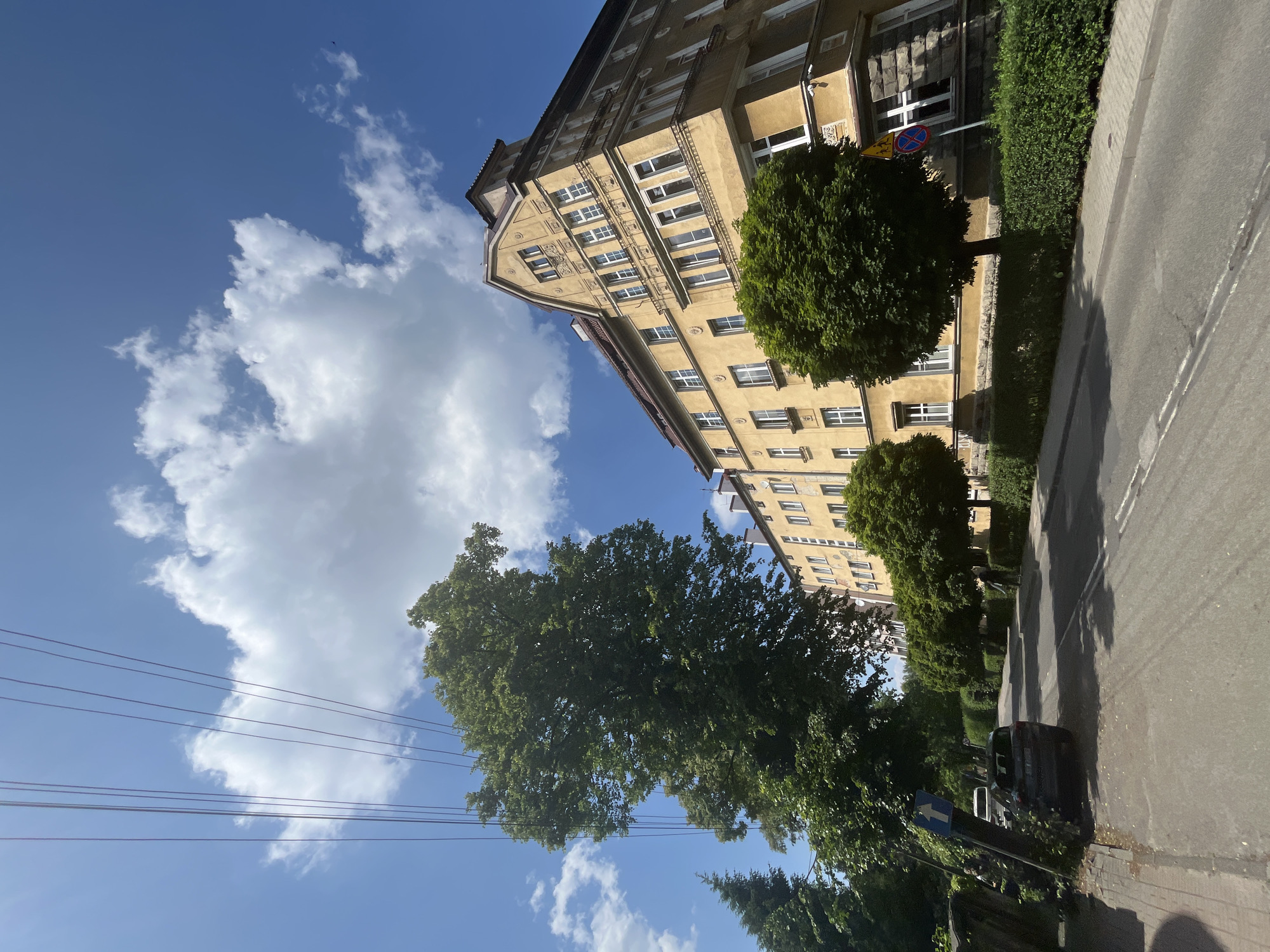 The image captures an urban or suburban street scene on a bright, sunny day, with a large building, trees, and a road under a blue sky.
A tall, multi-story building, likely an apartment complex, dominates the right side of the frame. It features a light yellow or beige facade with numerous white-framed windows and some decorative architectural elements, particularly visible on the detailed gable end at the top. The building's design suggests it is of an older construction.
Lining the street are several vibrant green trees with dense foliage, with one particularly large tree prominent in the foreground and mid-ground. Hedges run alongside a grey paved sidewalk. A dark-colored car is parked on the street, partially obscured by shadows and the surrounding greenery. The paved street features faint white markings.
Several street signs are visible: a triangular warning sign with a red border and yellow background, depicting a black symbol resembling a vehicle skidding or a crossroads; a circular sign with a red border and a blue diagonal line (likely indicating "No Stopping" or "No Waiting"); and a blue rectangular sign with a white arrow pointing upwards, possibly indicating a one-way street or direction. Thin power lines stretch across the upper left portion of the blue sky, which is dotted with scattered white cumulus clouds.
The scene depicts a calm daytime setting, likely morning or afternoon, given the bright sun and distinct shadows cast by the trees and building. No people are visible, and no specific activity is taking place. There is no legible text on any of the signs or the building.