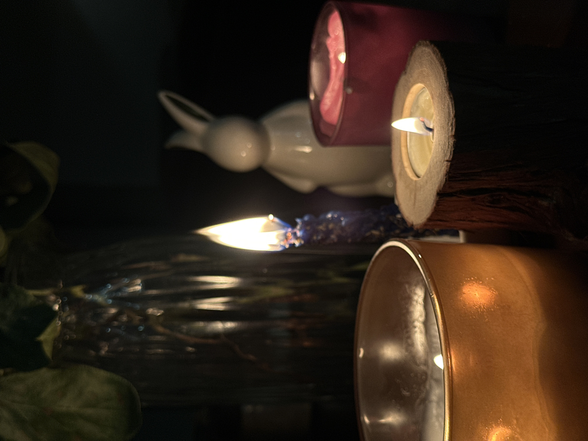 The image shows a dimly lit indoor scene featuring several decorative elements. Two lit candles are prominent, one in a wooden holder and another in a golden container. A pink cylindrical object is partially visible behind the wooden candle. A white ceramic bunny figurine is in the background, partially obscured by the foreground elements. In the lower left, there are hints of greenery, possibly a plant. The reflective surface beneath the candle flame suggests a glass or mirrored surface. The image appears to be taken in a dimly lit, possibly domestic setting. There is no information to indicate the location is Chaikovsky, Russia.