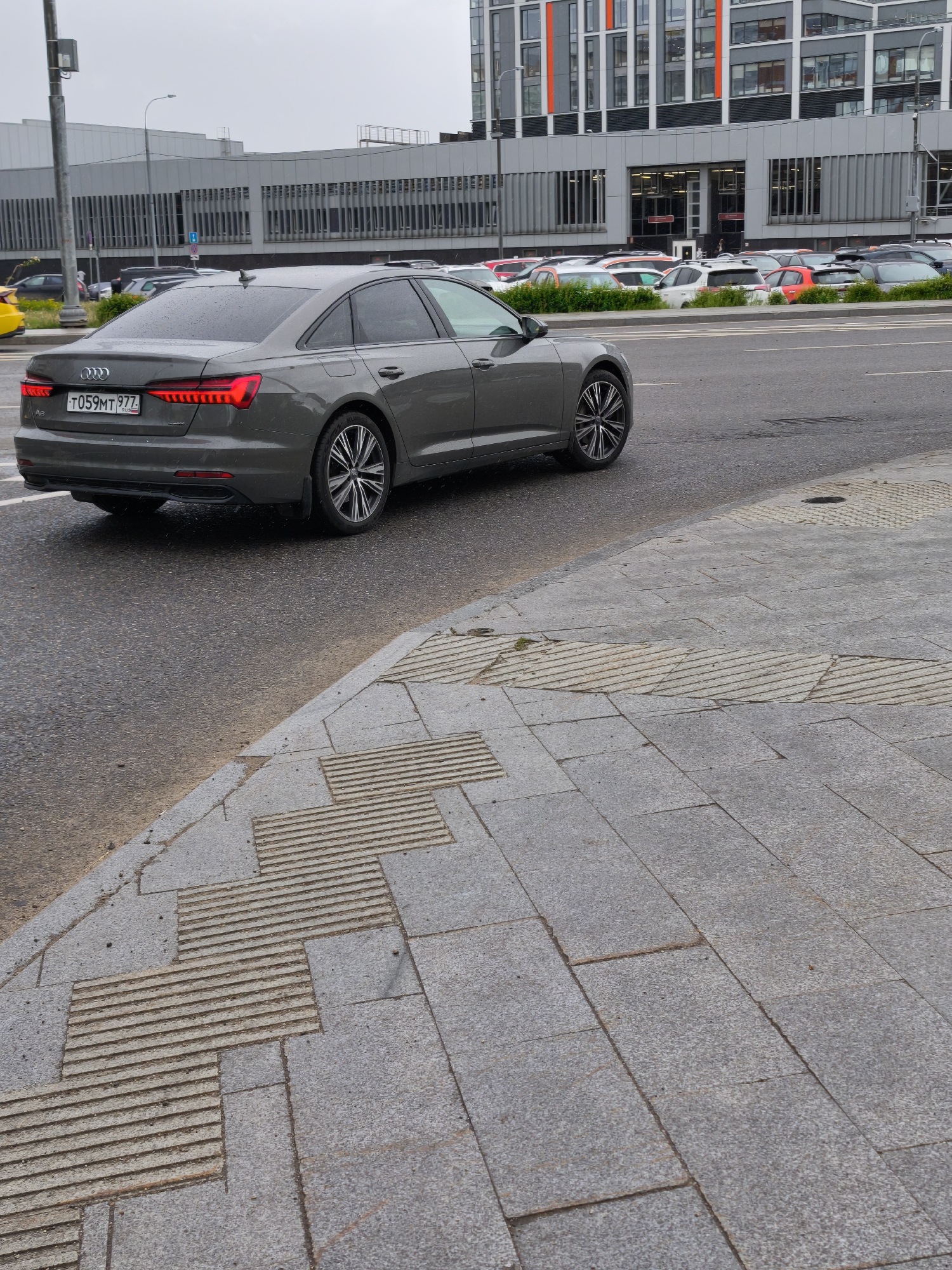 The image depicts a gray Audi A6 car with the license plate "T059MT 777" on a wet road in Moscow, Russia. The car is traveling away from the camera, and its rear lights are illuminated. In the background, there is a parking area with multiple cars and a modern building with gray and orange facade elements. There is also a gray pedestrian bridge or walkway above the road. The foreground shows a sidewalk paved with rectangular gray stones, some of which have textured surfaces. The overall environment suggests an urban setting on a rainy day.