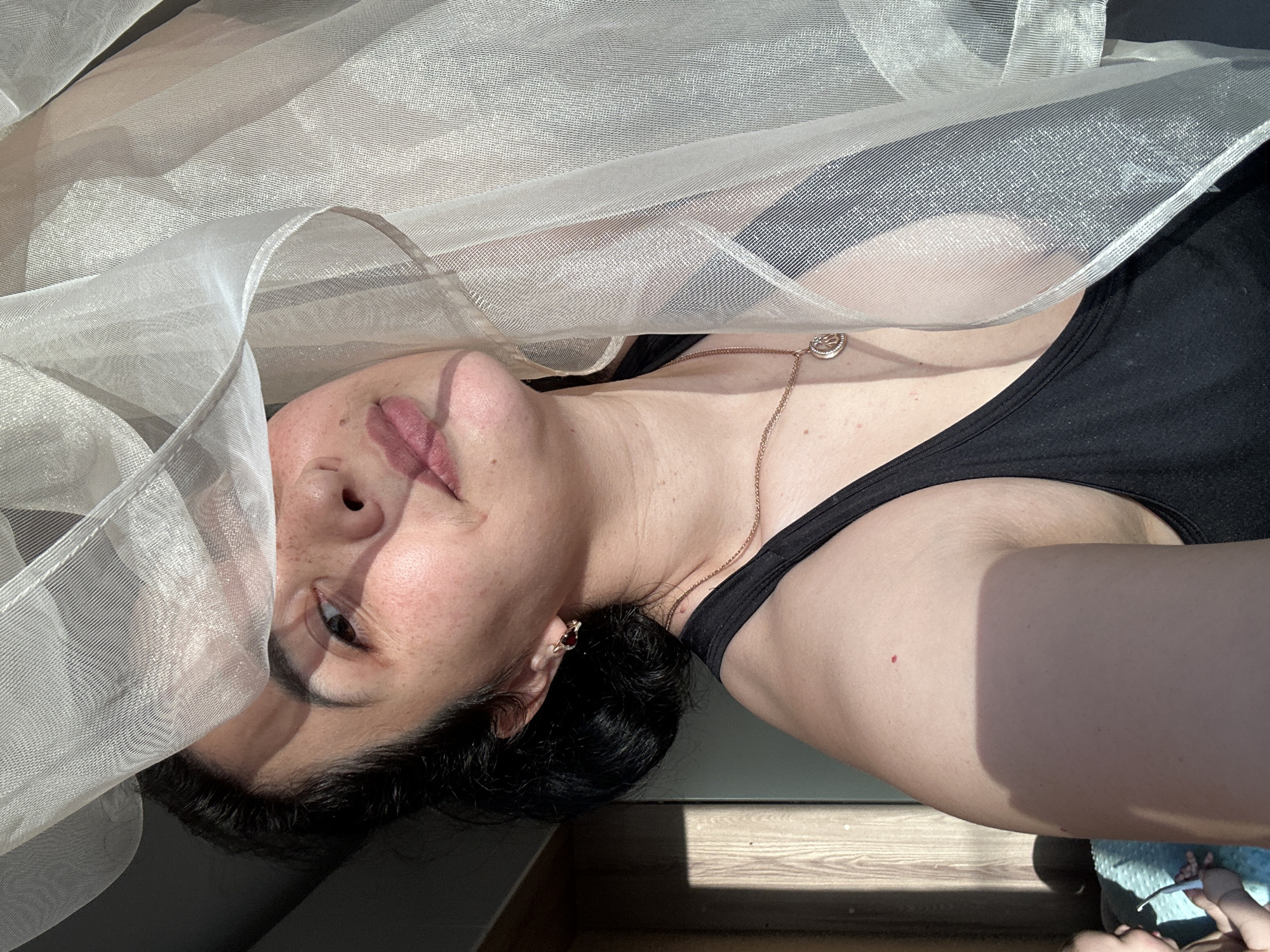 A woman is captured in a close-up, waist-up, indoor portrait. She is positioned in the center, partially obscured by a sheer, light-colored curtain that drapes across the upper portion of the frame, creating a play of light and shadow on her face. The woman has dark hair pulled back, and her head is tilted slightly towards the camera, with her right eye visible. She is wearing a black tank top and a delicate gold chain with a circular pendant. Her skin shows freckles and is illuminated by sunlight casting a warm glow and distinct shadows. To the left of the woman, a portion of a wooden-paneled wall is visible, with a distinct shadow line indicating sunlight coming from a window. In the lower left corner, a small section of a baby is visible, with a hand reaching out towards a blue object. The overall impression is one of a casual, sunlit moment.