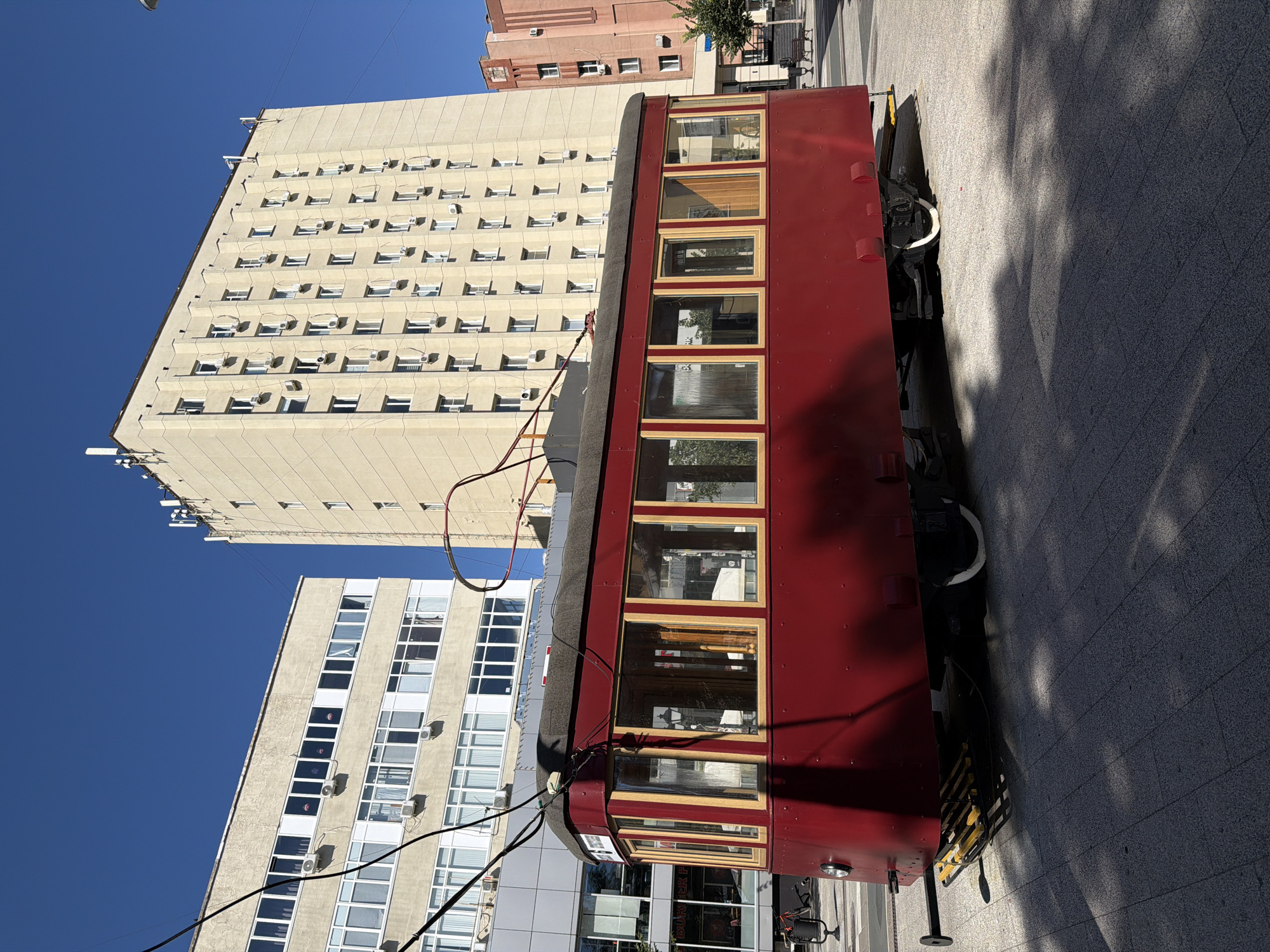 An old, dark red streetcar sits on display on a paved plaza in Saratov, Russia. The streetcar, which appears to be made of wood and metal, has large windows with wooden frames. It is situated in front of several modern buildings, including a tall, light-colored apartment building with many windows and air conditioning units, and a lower, white building with similar architectural features. The sky is clear and blue. Shadows from trees are cast across the plaza and the streetcar, suggesting it is a sunny day. The ground is paved with grey interlocking stones. There are no people visible in the image. The streetcar is likely a historical exhibit or monument in the city.
