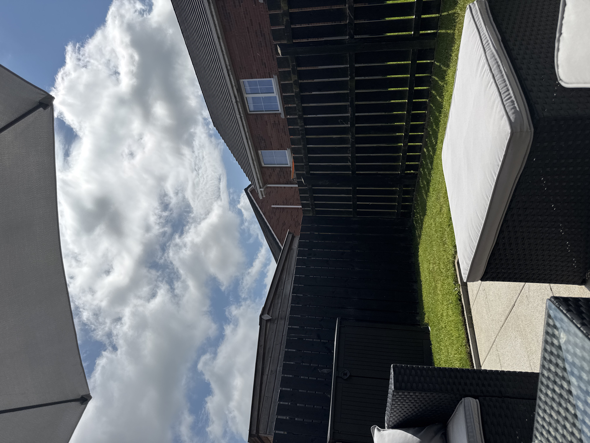 The image depicts a residential backyard in Craigavon, United Kingdom, on a sunny day with scattered clouds. The foreground features outdoor furniture, including a black rattan sofa with grey cushions and a matching footstool also with grey cushions. A glass-top table is partially visible in the lower left corner.
Behind the furniture, a neatly mowed green lawn meets a paved patio area. A tall, dark wooden fence runs across the midground, separating the backyard from neighboring properties. Beyond the fence, a portion of a wooden shed is visible on the left, and a brick house with a tiled roof and white-framed windows is seen on the right. The sky above is a bright blue, populated by fluffy white clouds, and a large, light grey umbrella is positioned in the upper left of the frame, casting a shadow. The scene conveys a typical domestic outdoor setting.
